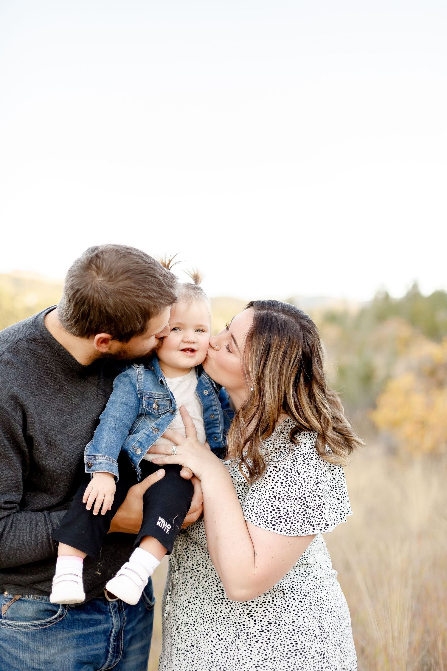 Young parents each kissing the cheeks of their smiling toddler daughter in an autumn field.