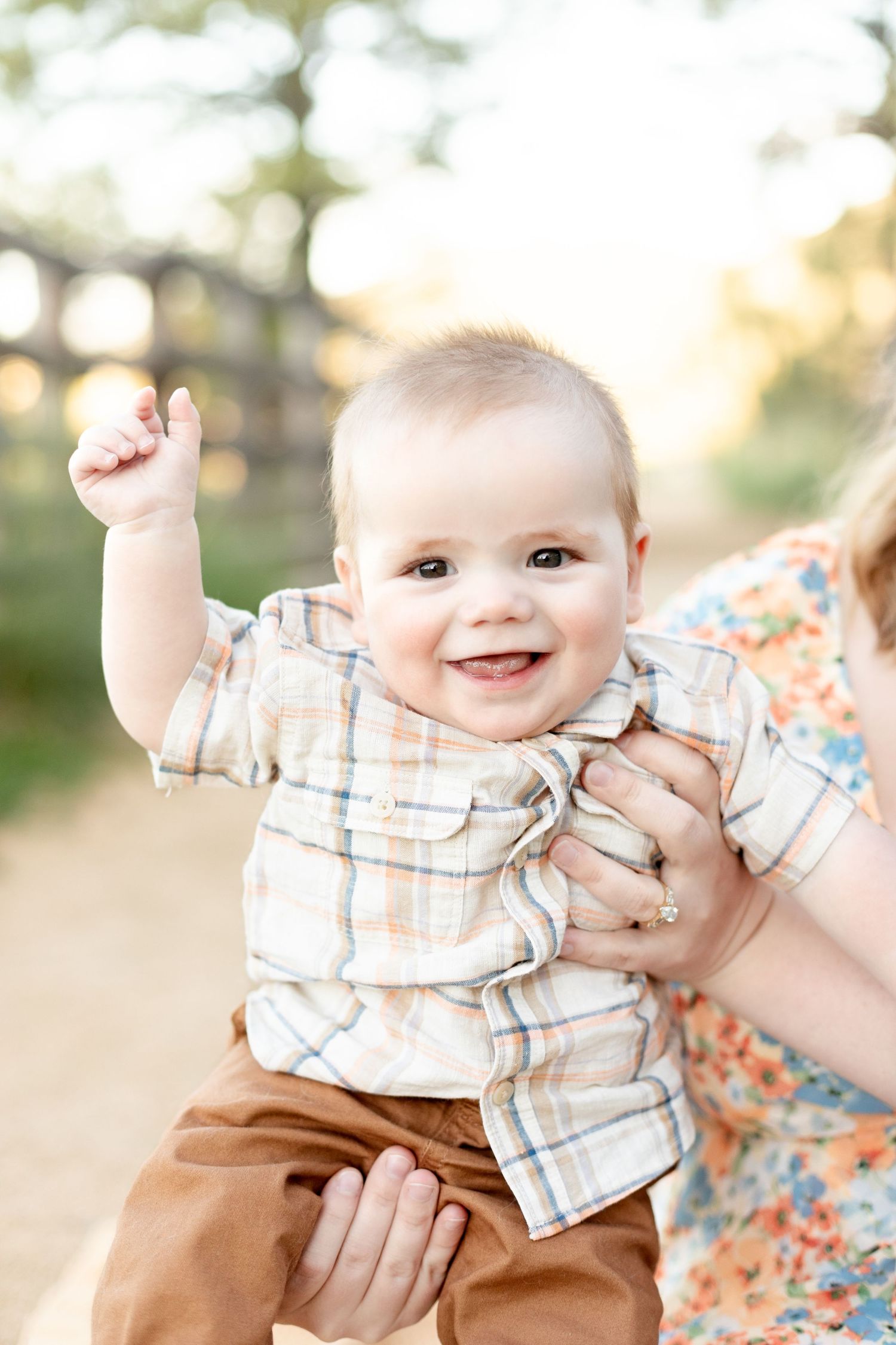 Happy smiling baby in plaid shirt raising fist outdoors during family photo session.