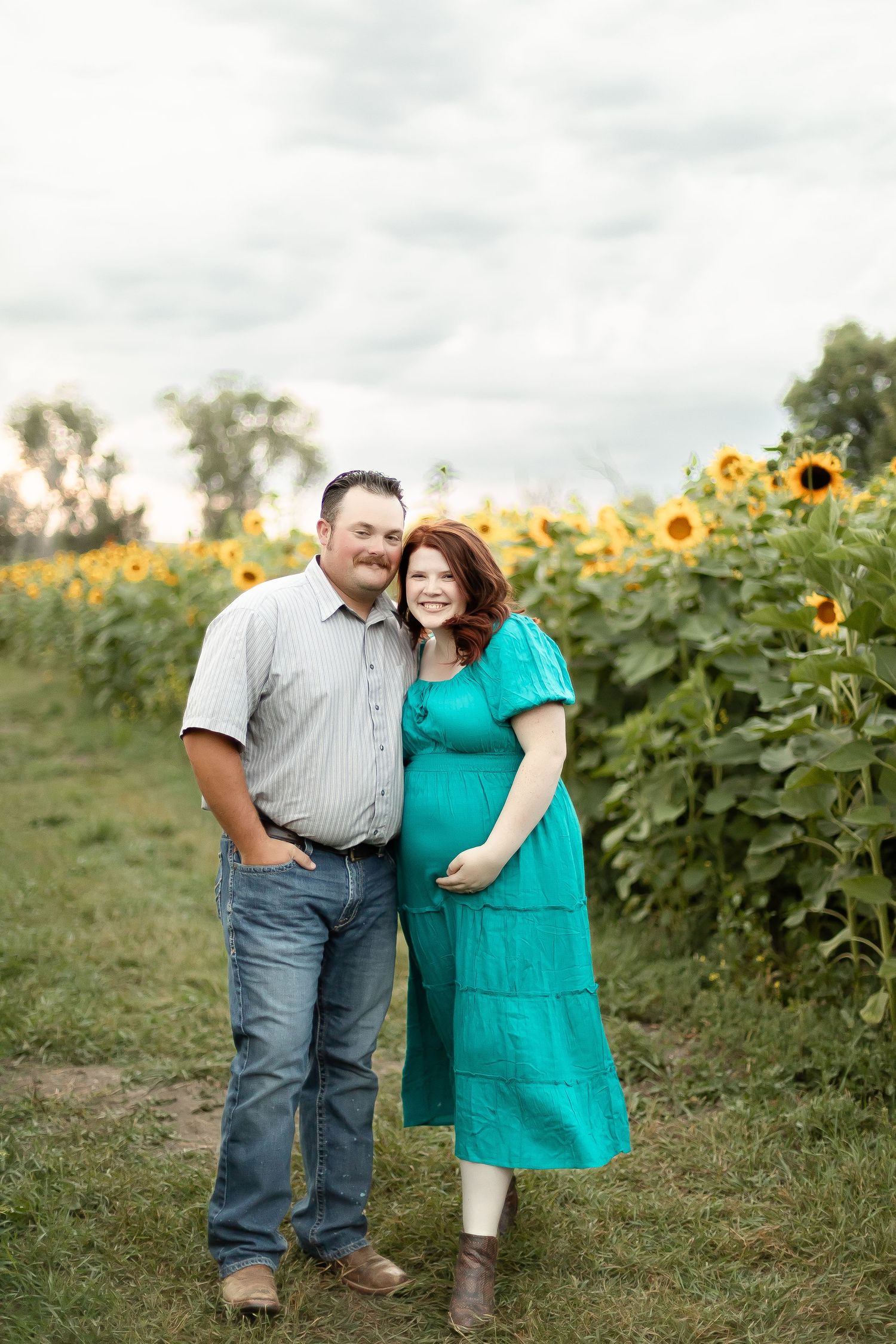 Couple smiling together in front of a sunflower field, woman in teal maternity dress showing baby bump.