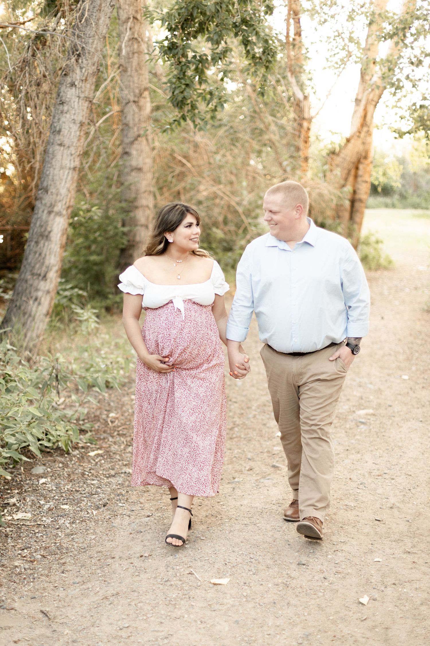 Happy pregnant couple walking hand in hand on a tree-lined path during a warm golden hour maternity session.