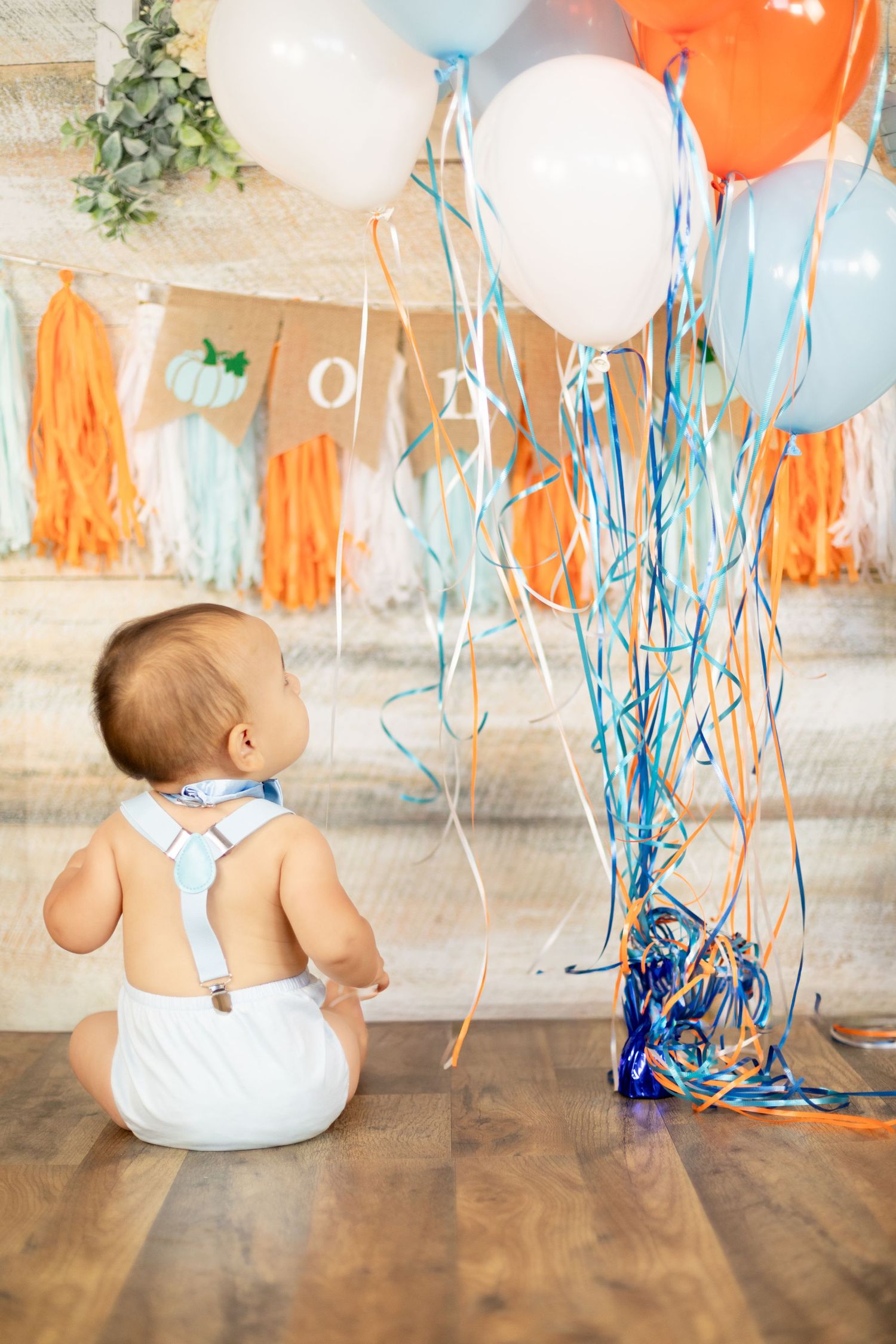 Baby boy in white diaper with suspenders sitting facing colorful balloons and streamers at first birthday party.