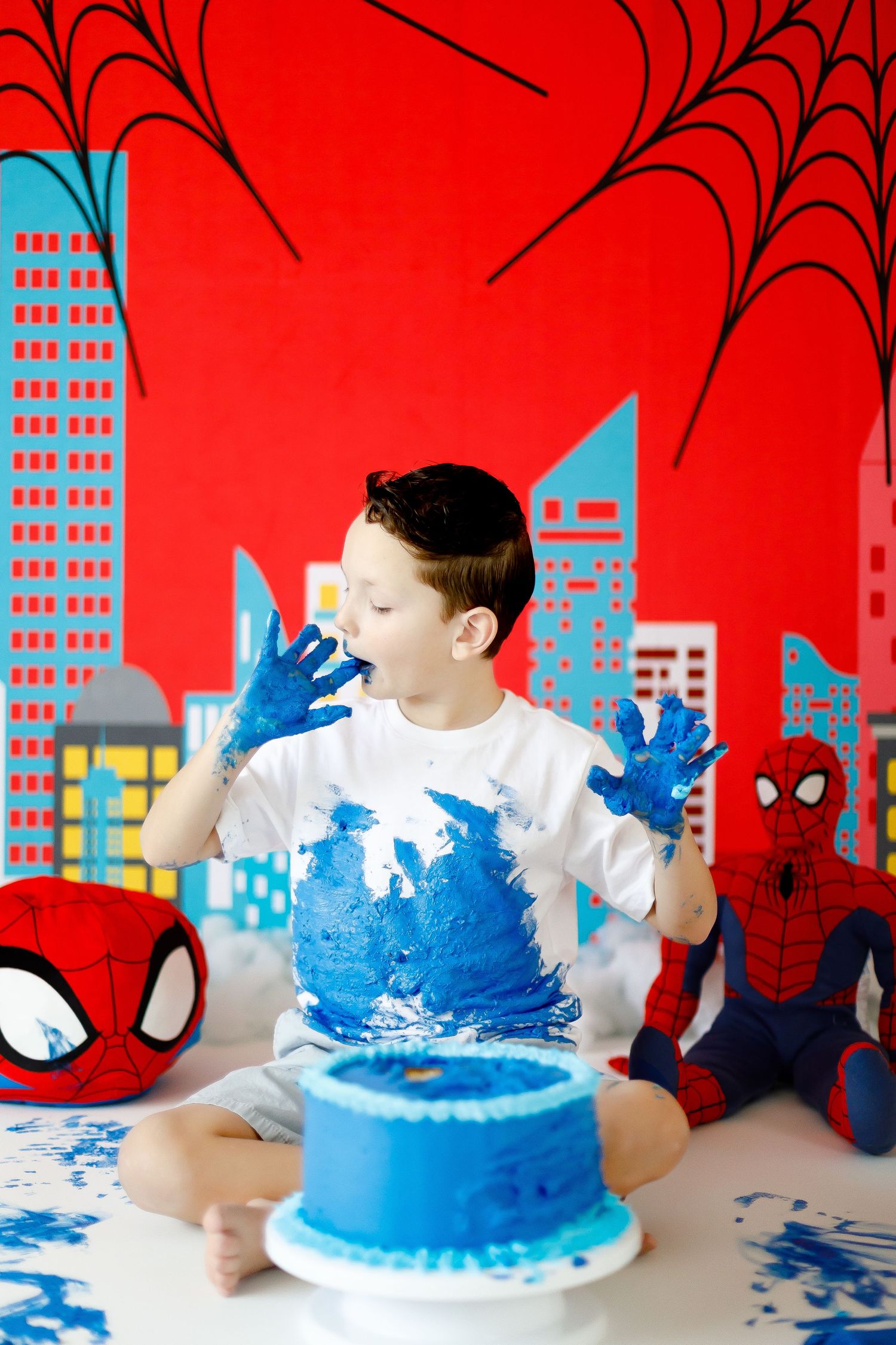 A boy covered in blue frosting licks his hands after smashing a blue birthday cake.