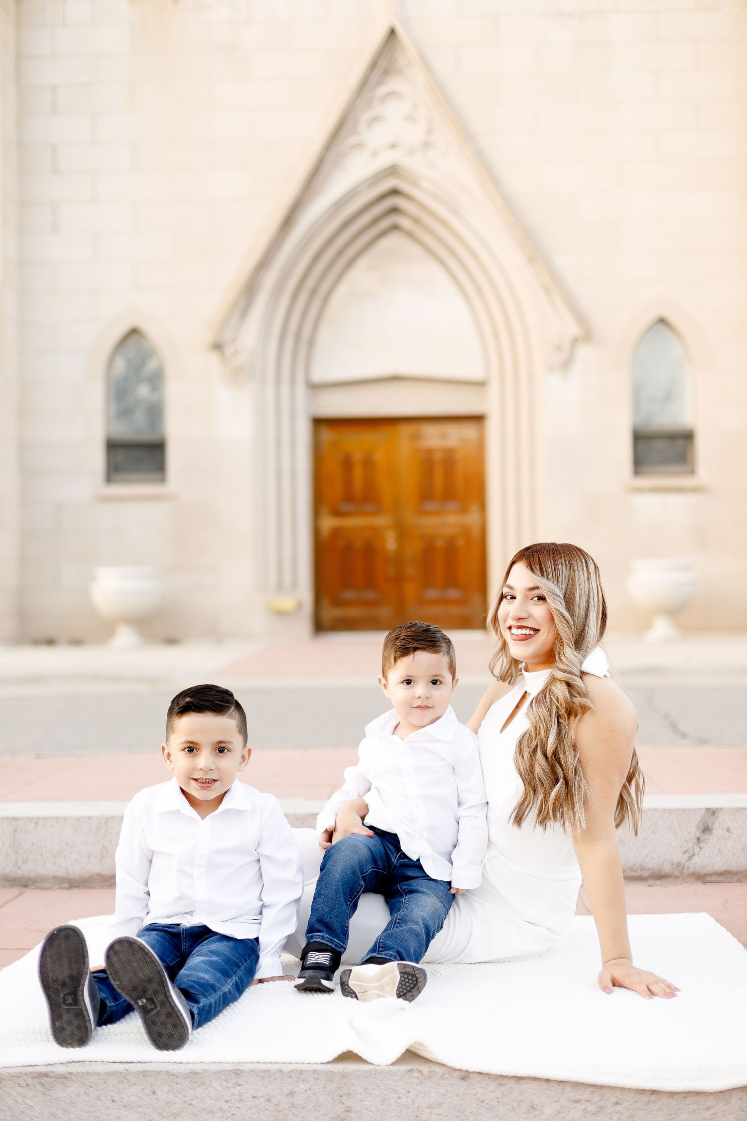 Mother in white dress sitting with two young boys in front of church doors.