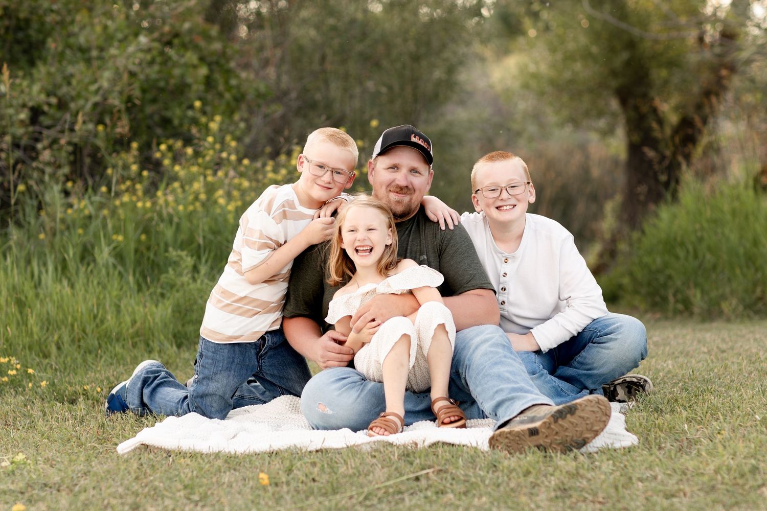 A father sits on a blanket in a field with his three young children, all smiling outdoors.