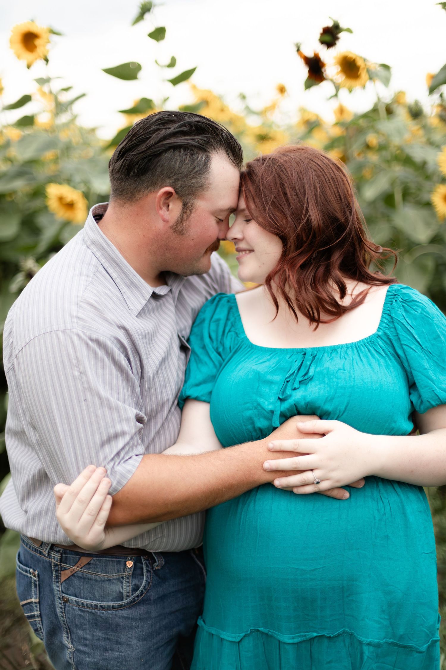 Pregnant woman in teal dress and partner share an intimate moment in a vibrant sunflower field.
