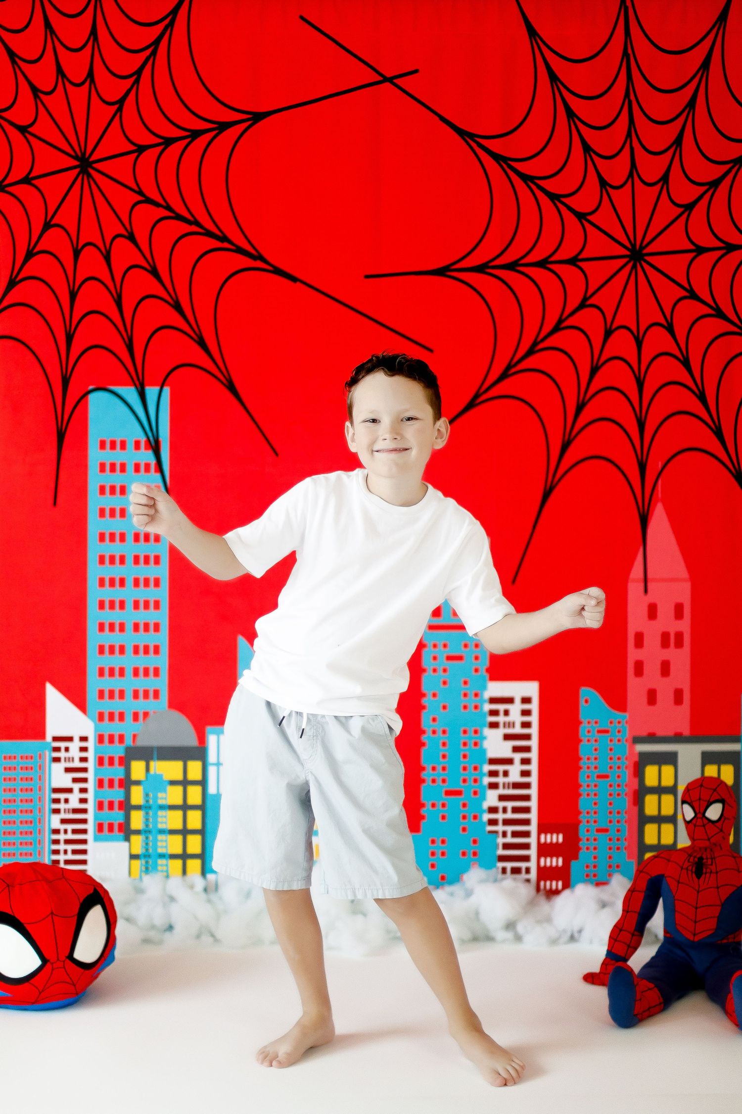 A boy strikes a Spider-Man pose in front of a colorful red cityscape superhero birthday backdrop.