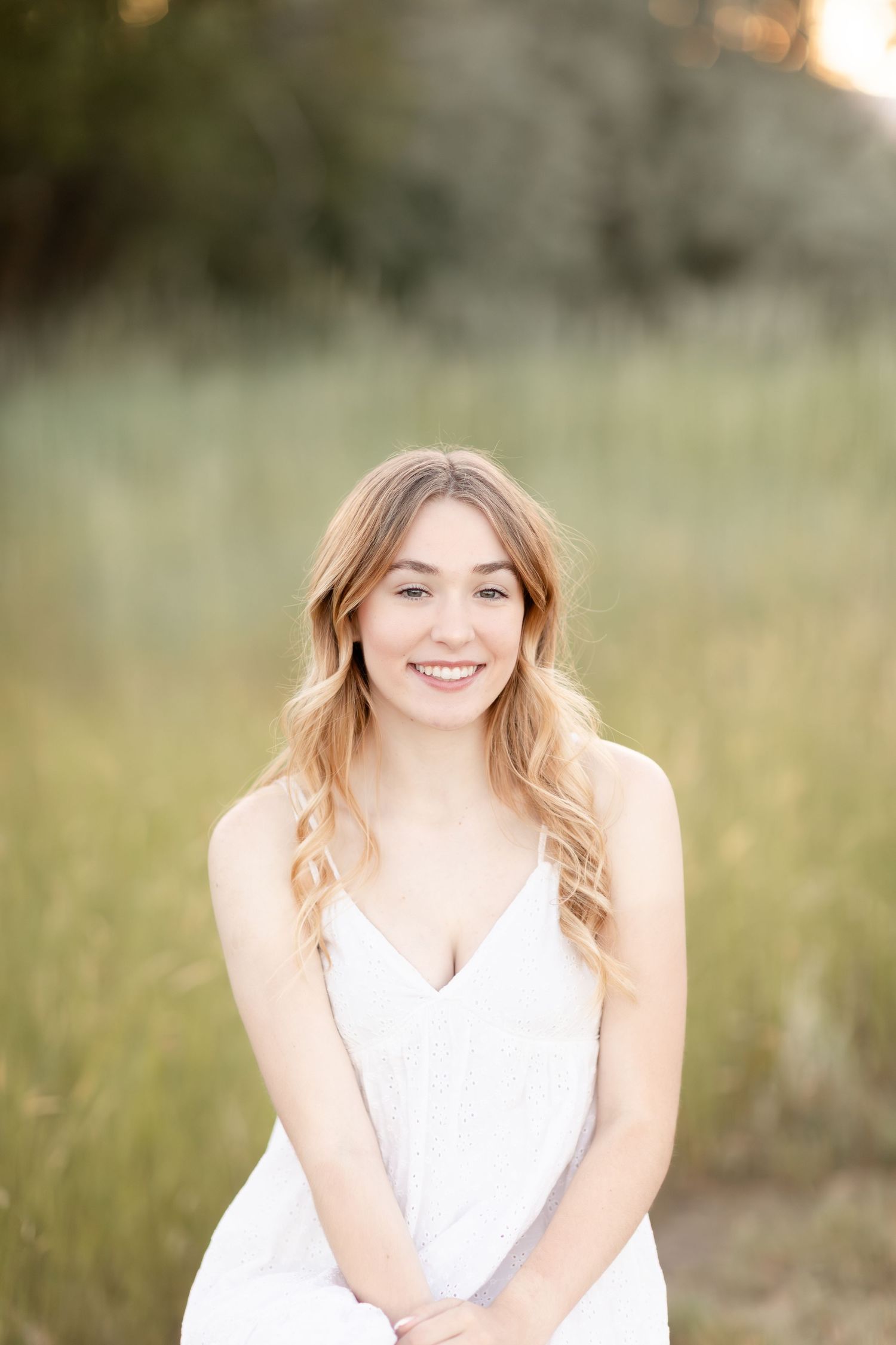 Young woman with long wavy blonde hair smiling outdoors in a white dress against a soft green meadow background.