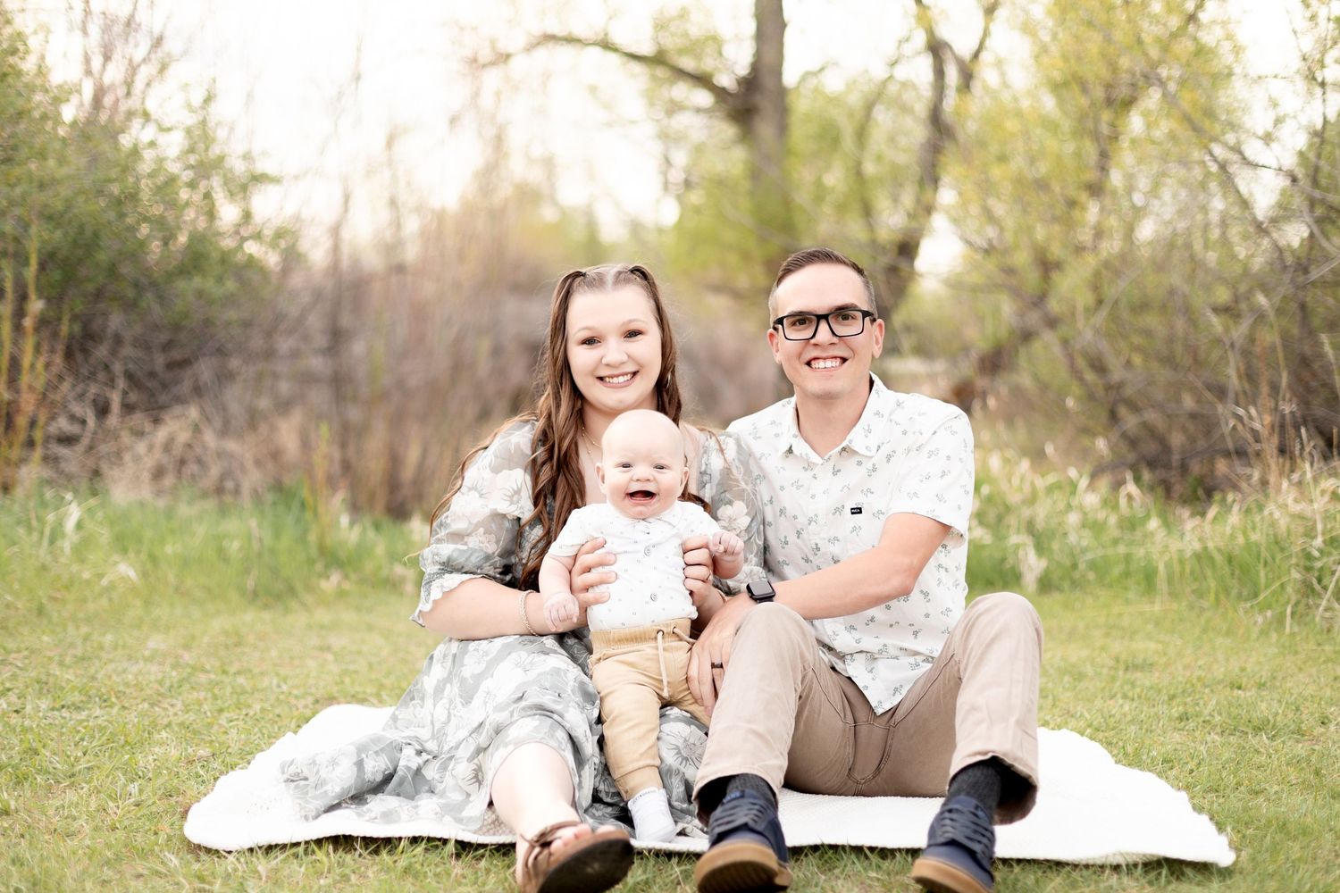 Happy young couple sitting on blanket in grass with laughing baby between them.