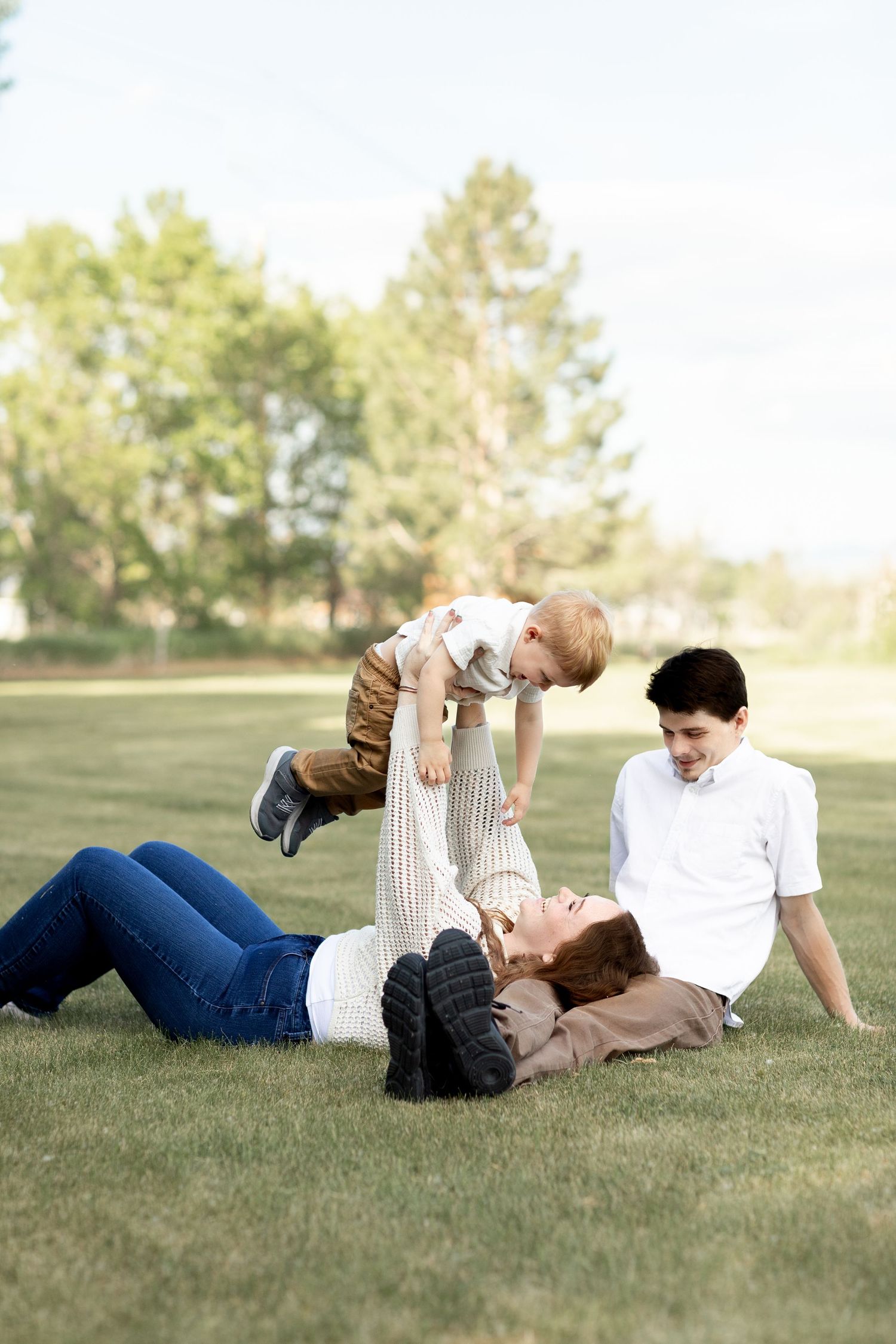 Young family playing on green grass as mother lifts toddler while father watches and smiles.