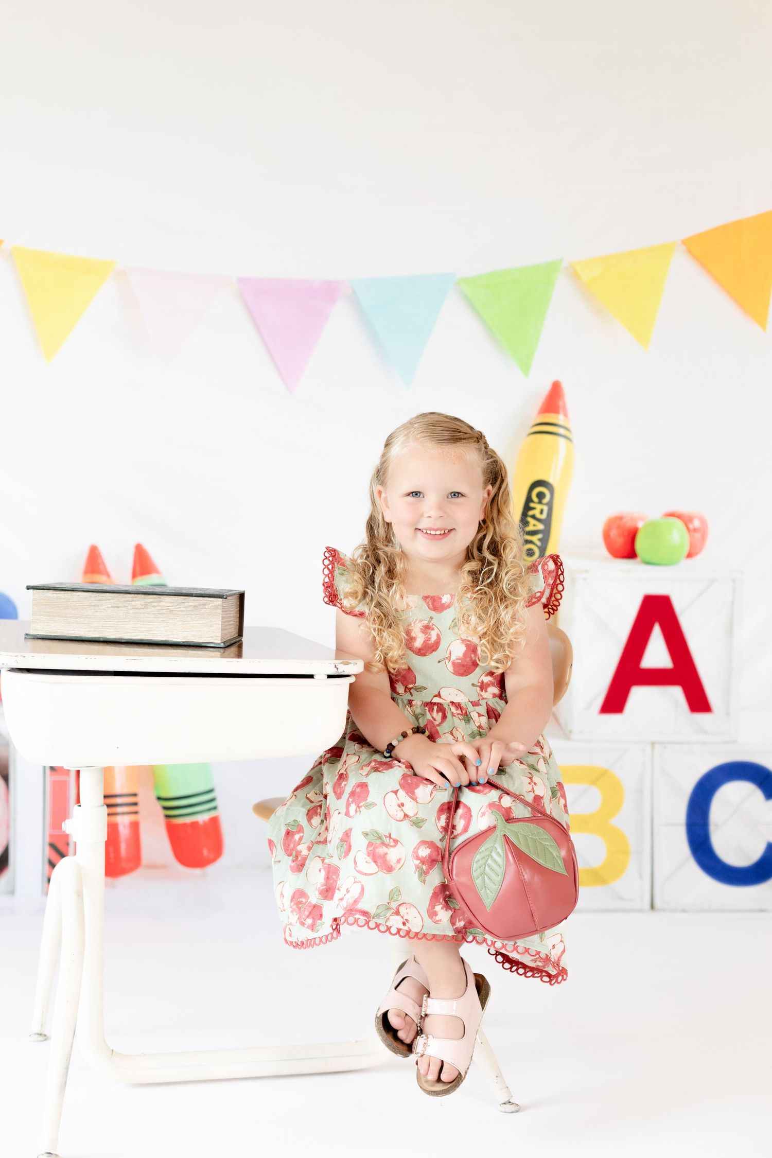 Young girl in floral dress holding giant crayon sits at school desk with ABC blocks and colorful bunting backdrop.