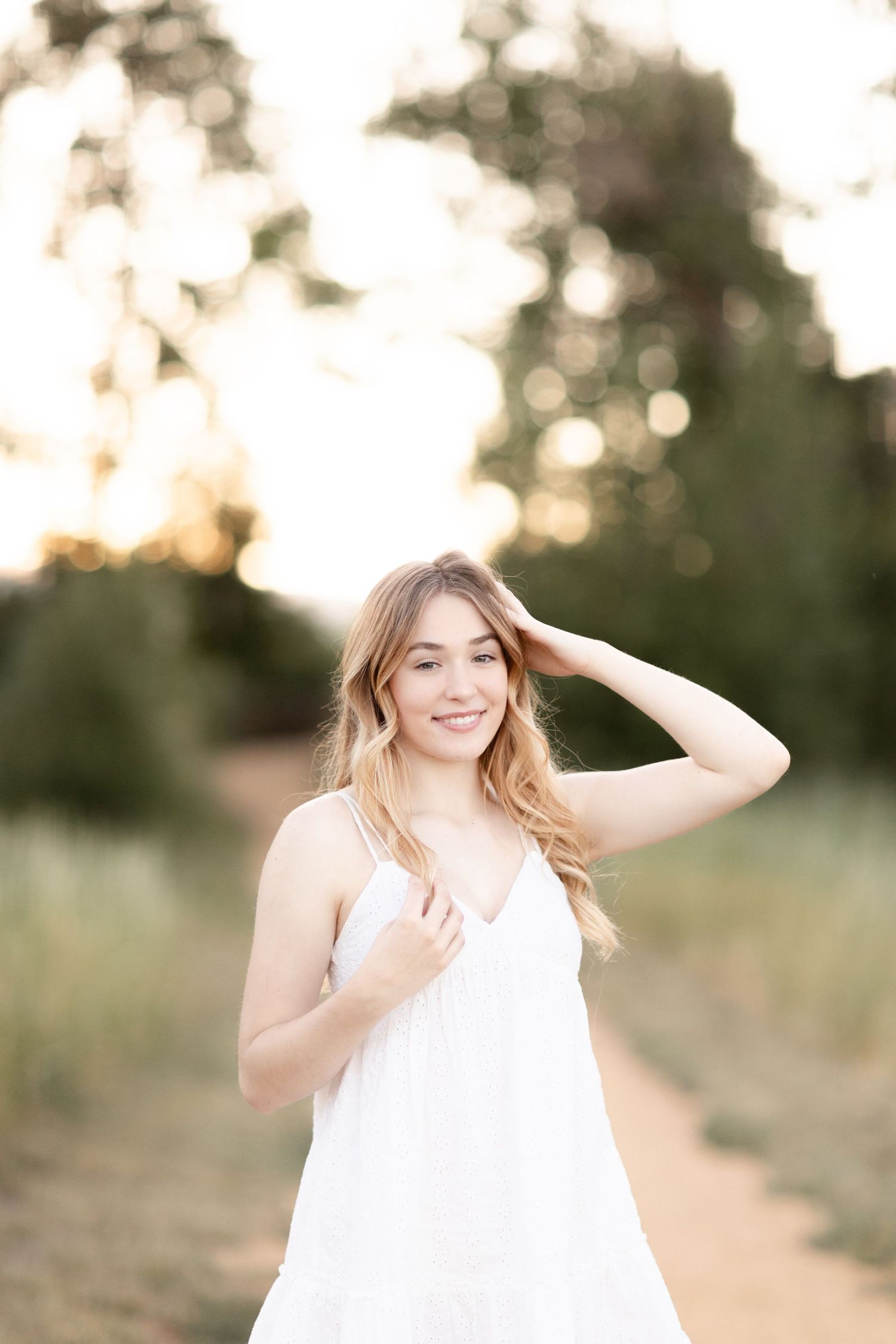 Young blonde woman in white sundress smiling outdoors on a path surrounded by lush green trees in golden light.