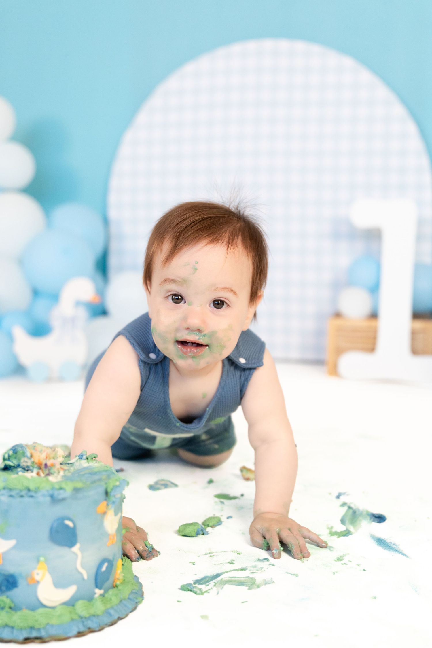 Baby boy with frosting on face crawling beside duck-themed blue smash cake at first birthday shoot.