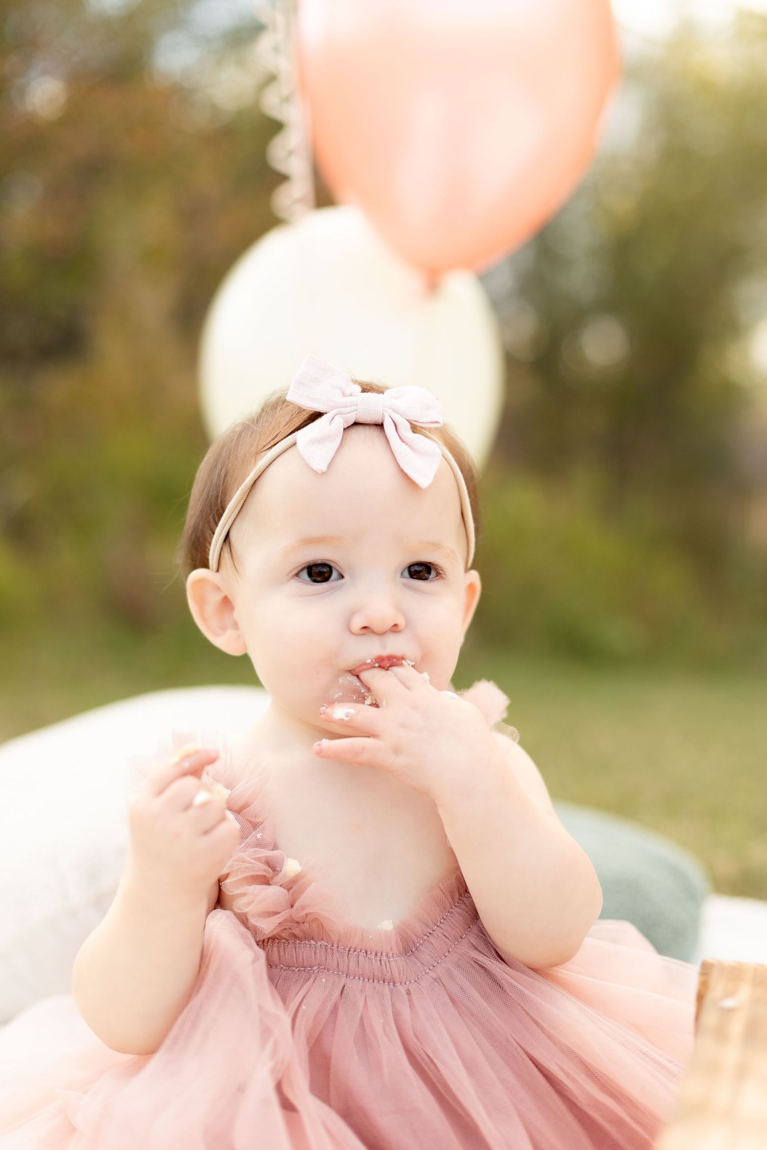Adorable baby girl in pink tutu dress with bow headband smashing birthday cake at outdoor party with balloons.