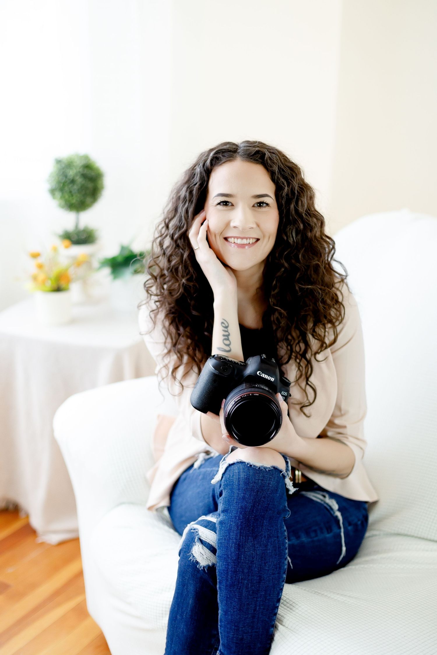 Curly-haired female photographer smiling on white sofa holding Canon camera.