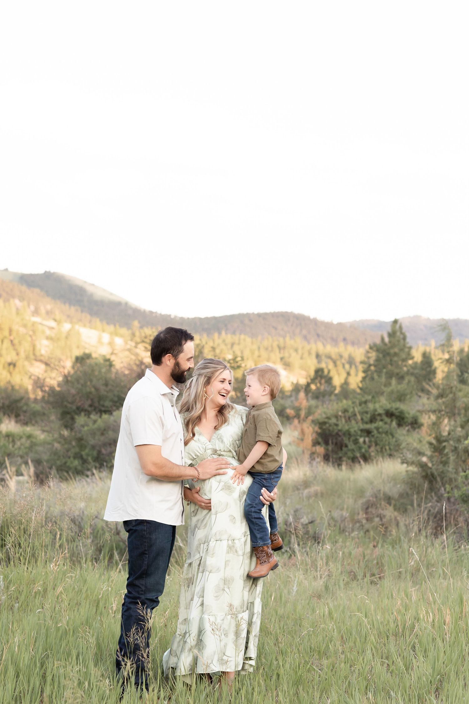 Happy expecting couple with toddler son standing in lush mountain meadow at sunset.
