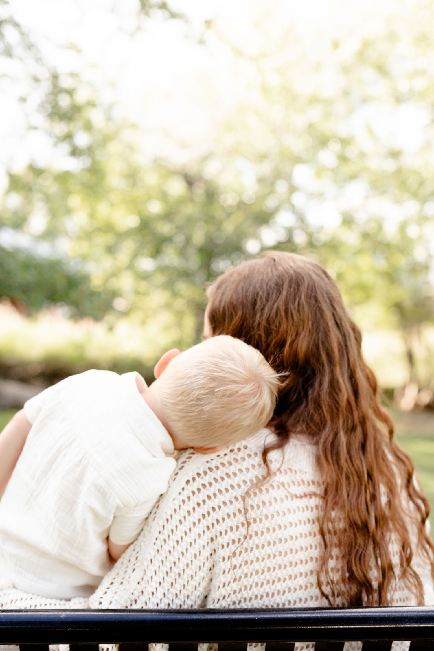 Tender moment of toddler snuggling against mothers shoulder while sitting on a park bench.