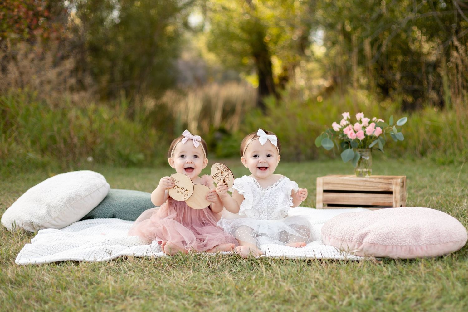 Twin baby girls in pink and white dresses smiling together outdoors at first birthday photo session.