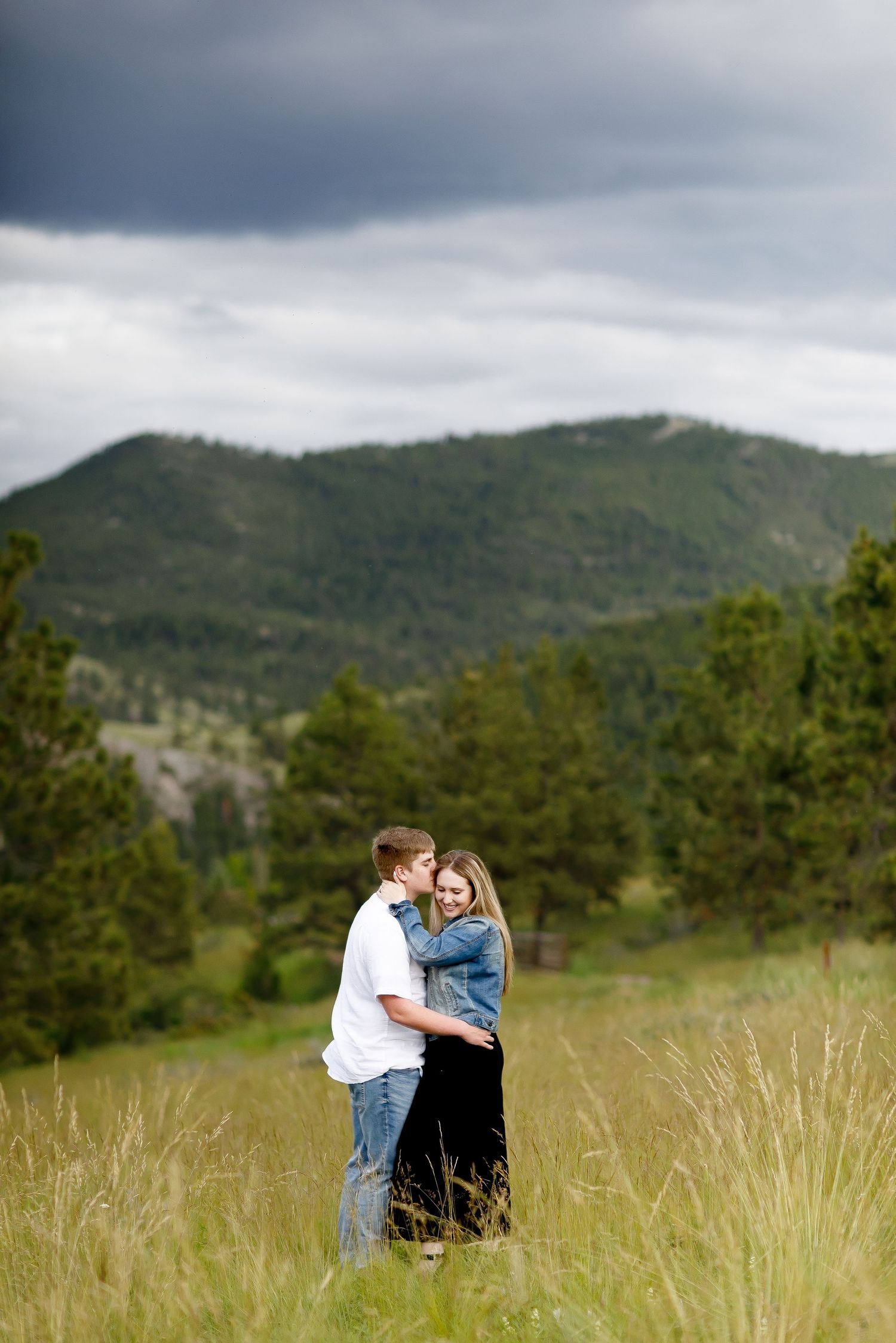 Couple embracing and kissing in a grassy mountain field under dramatic cloudy skies.