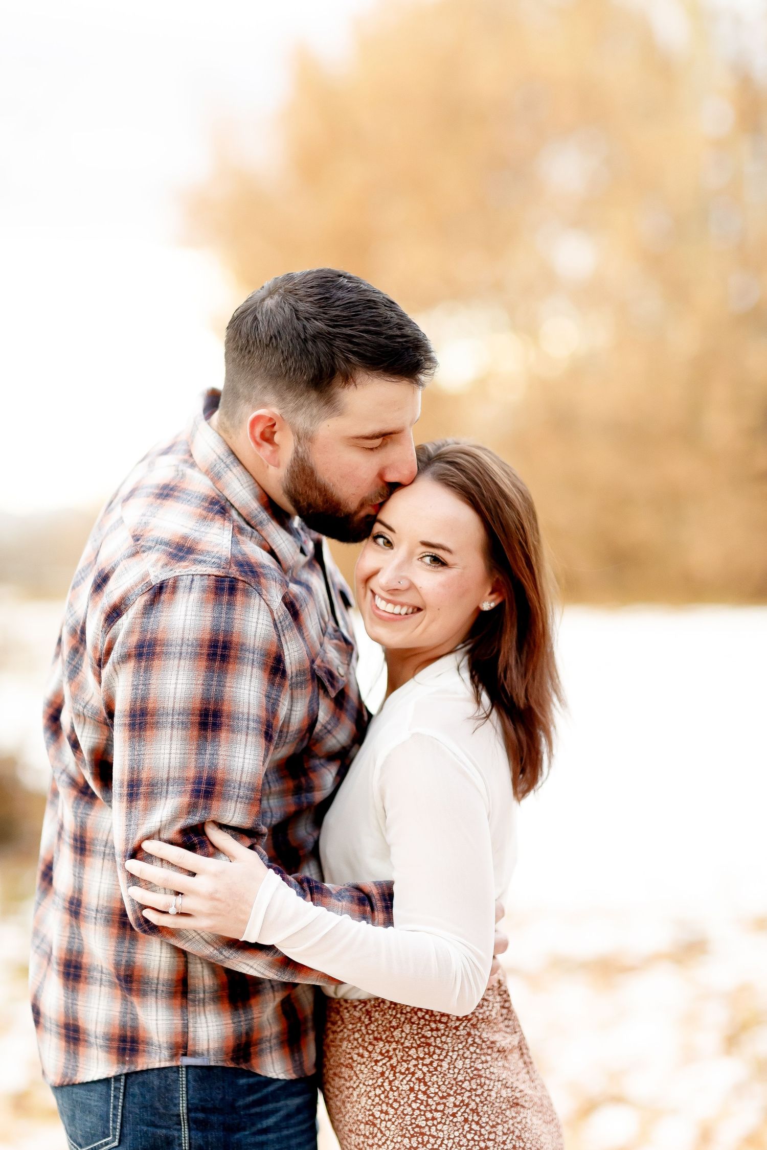 Man kissing woman on the cheek as she smiles warmly in a sunny autumn setting.
