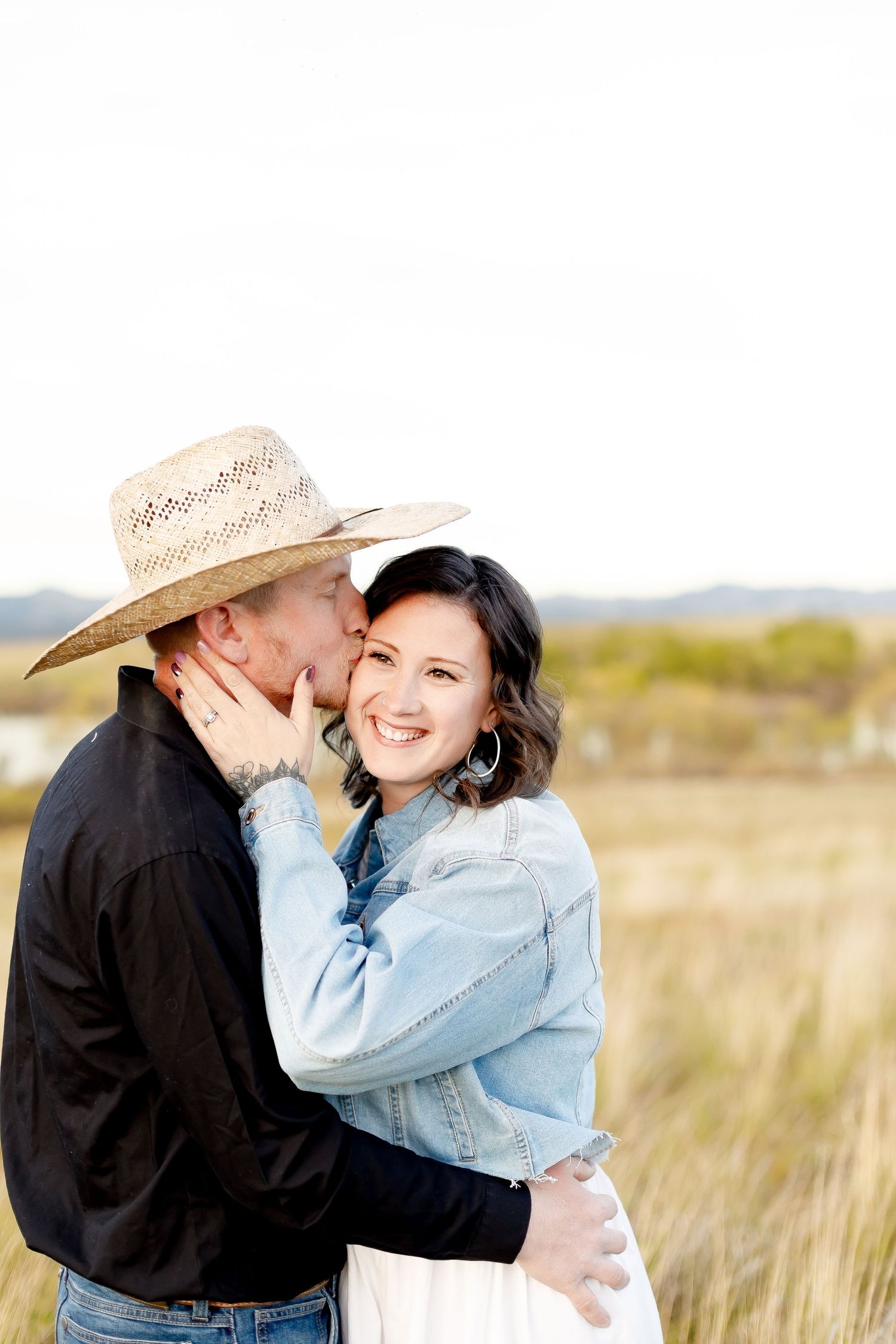 Man in straw cowboy hat kissing woman on cheek as she smiles in sunlit golden grass field.