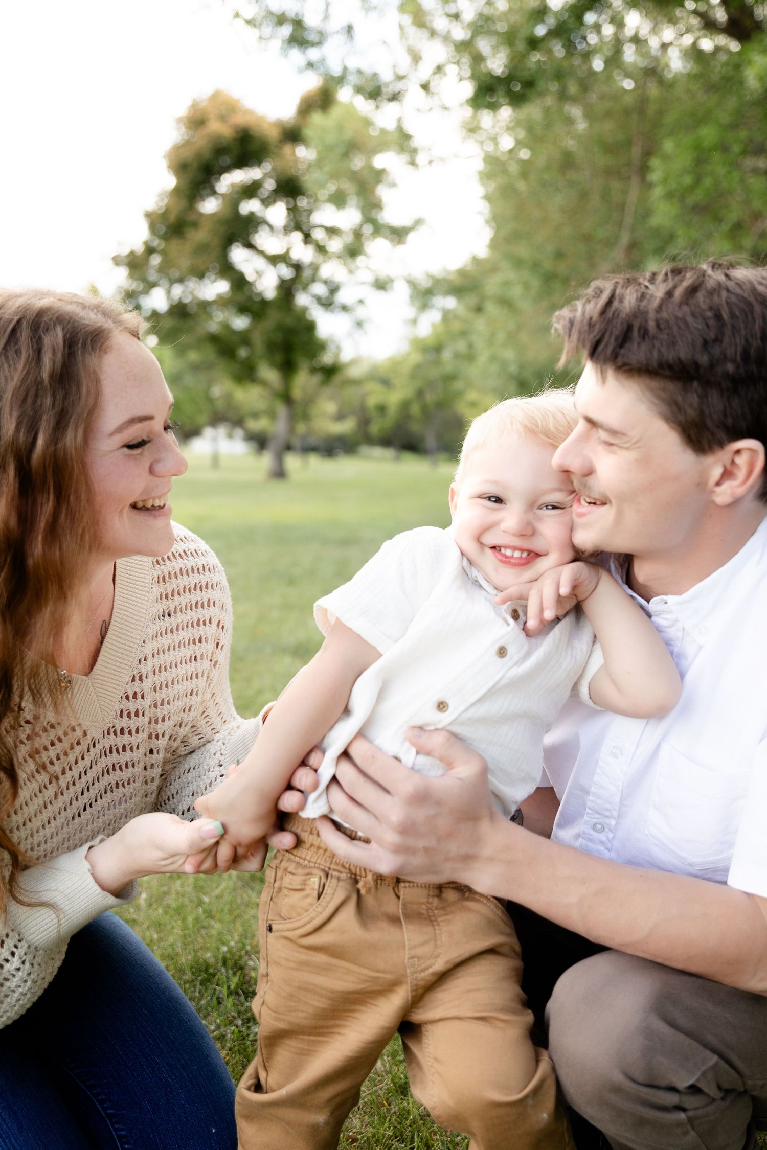 Happy young family with toddler son laughing together outdoors in a sunny green park.