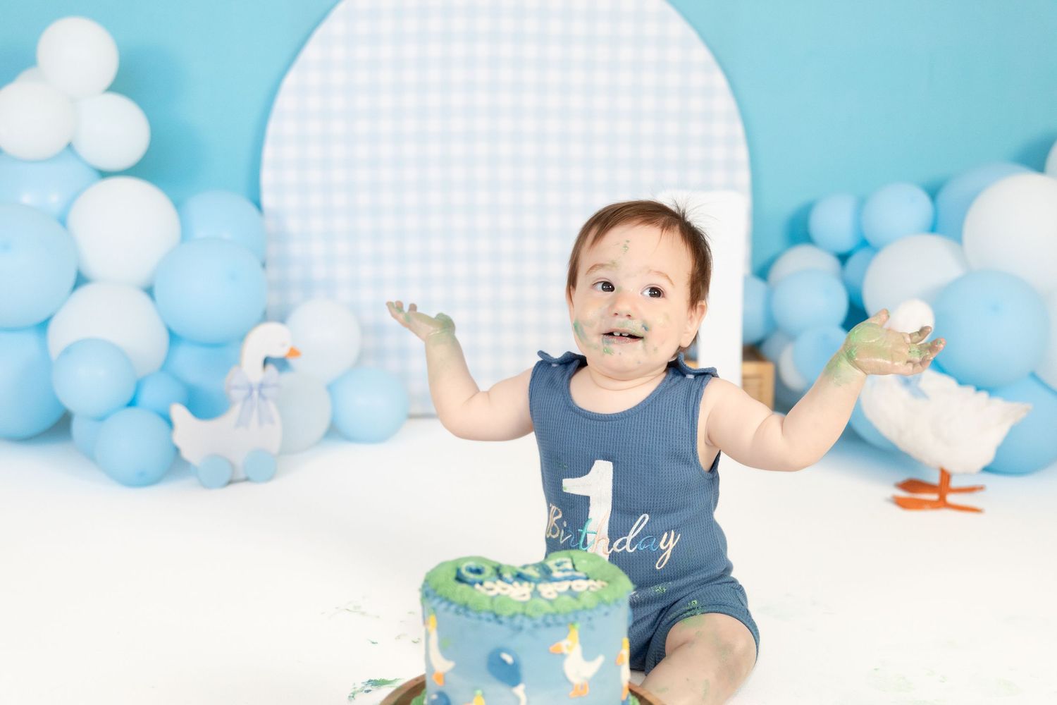 Laughing baby boy claps frosting-covered hands at first birthday cake smash with blue balloon backdrop.