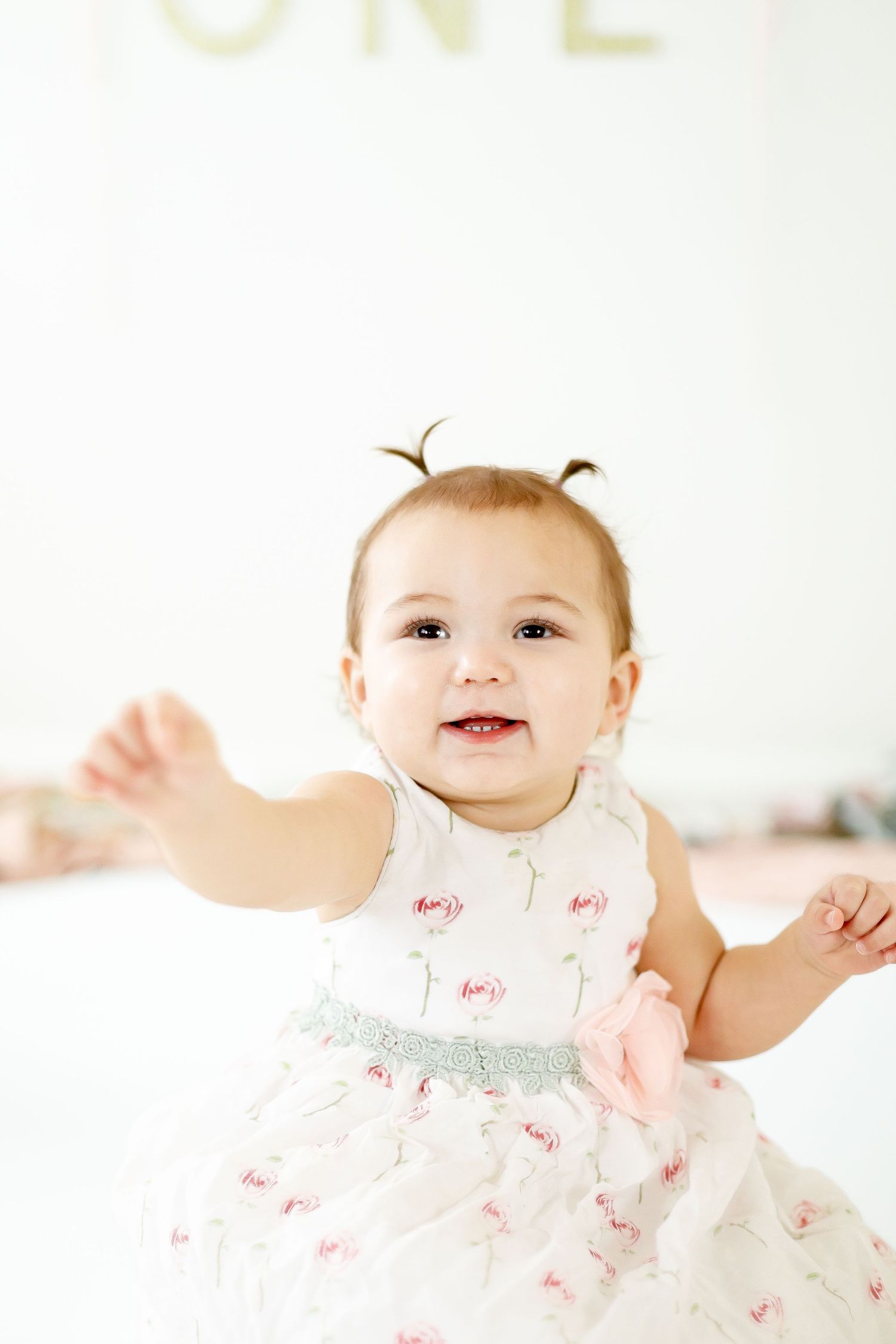 Adorable one year old baby girl in floral dress with pigtails smiling and reaching arms out.