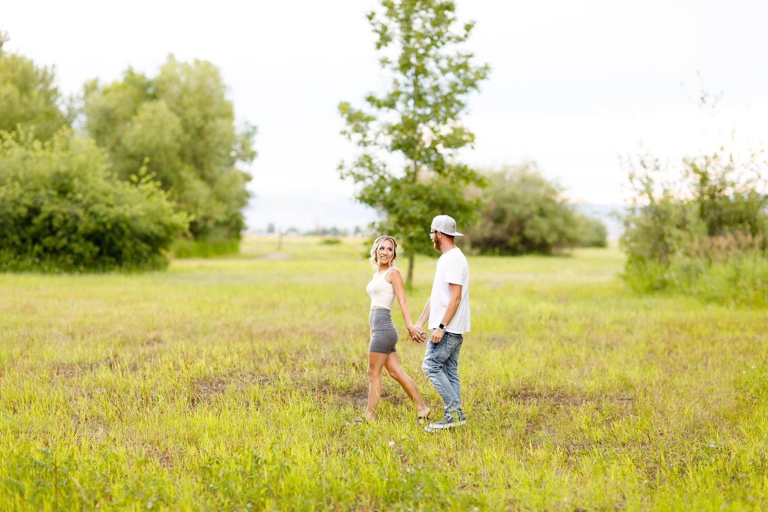 Happy couple holding hands and walking through a green meadow with trees in the background during engagement photos.