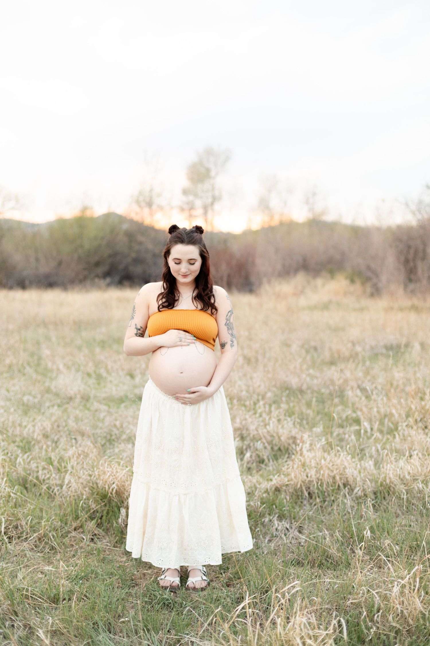 Pregnant woman in orange crop top and white lace skirt cradles her belly in a grassy field.