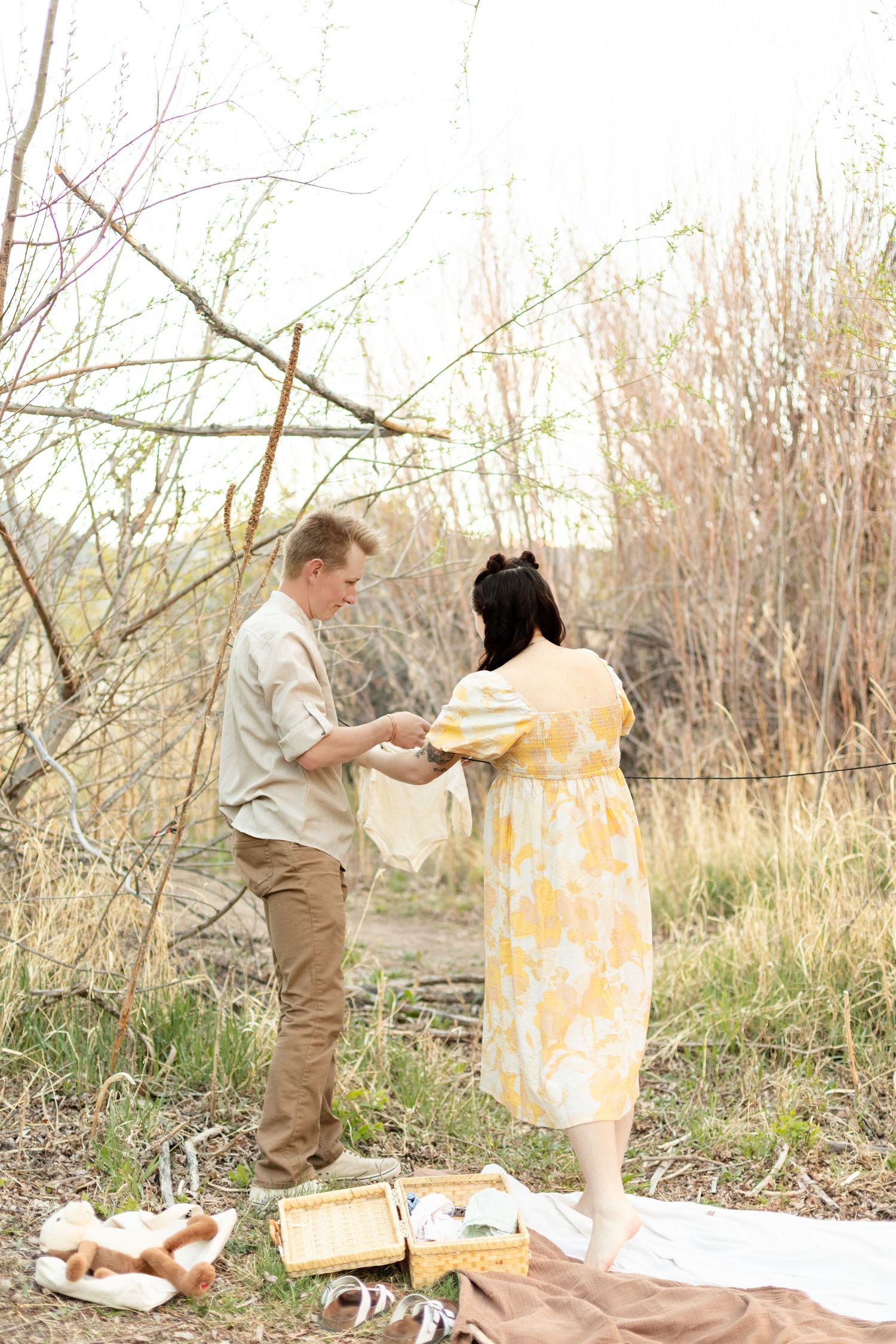 Expecting couple examines baby clothing outdoors on blanket surrounded by baby items near bare winter trees.