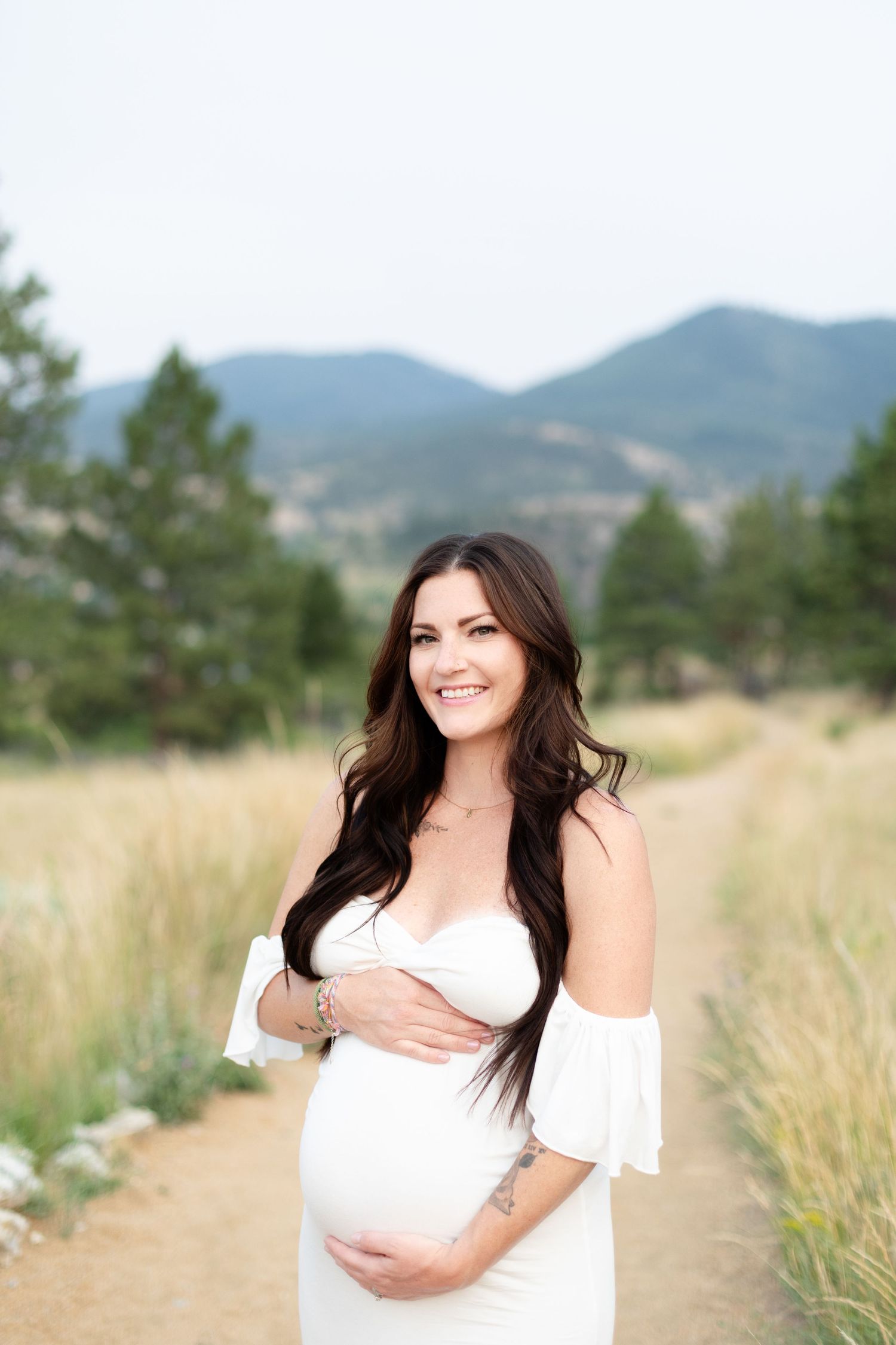 Smiling pregnant woman in white off-shoulder dress holding baby bump on mountain trail.