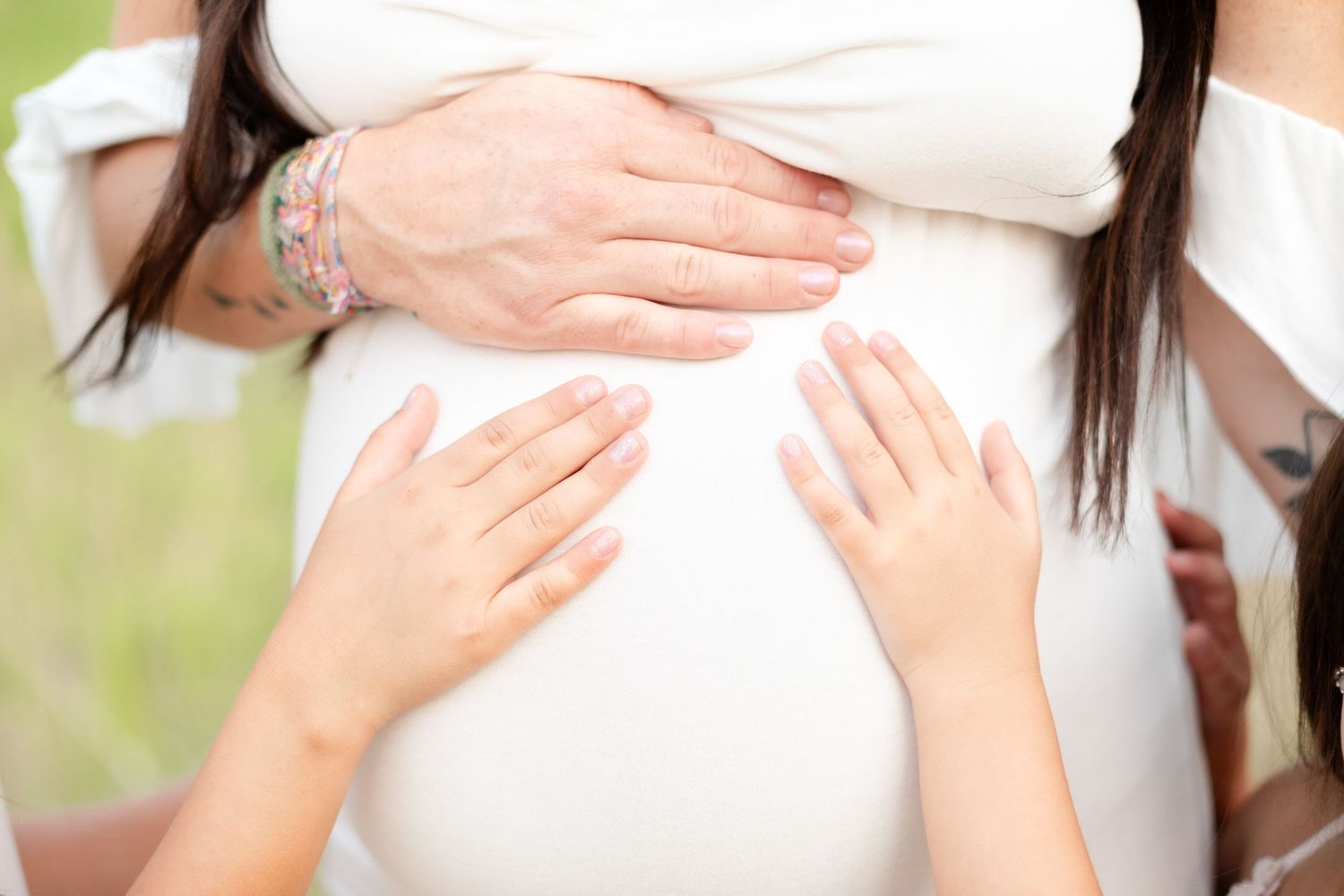 Close-up of pregnant belly with child hands touching white maternity dress.