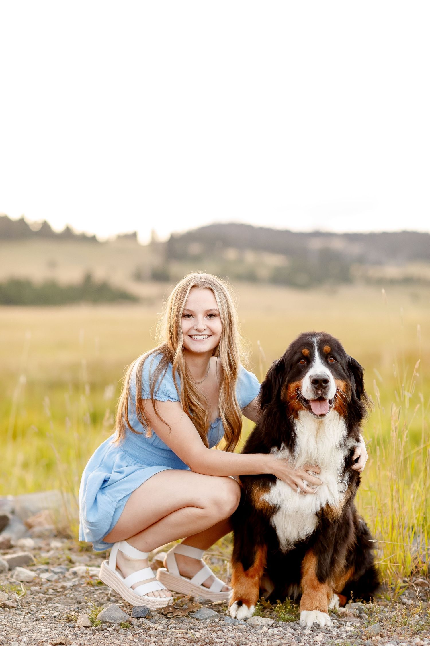 Smiling teenage girl in blue dress crouching beside a Bernese Mountain Dog in a grassy field.