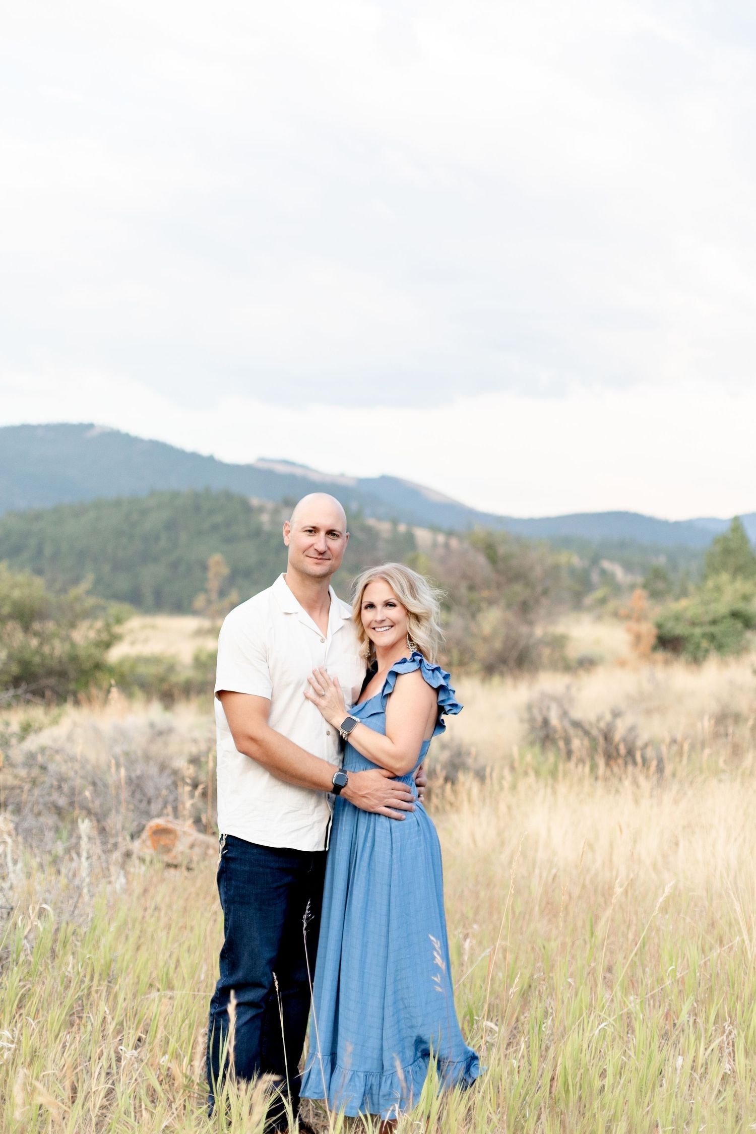 Couple embracing in a golden meadow with mountain views, woman in blue dress, man in white shirt.