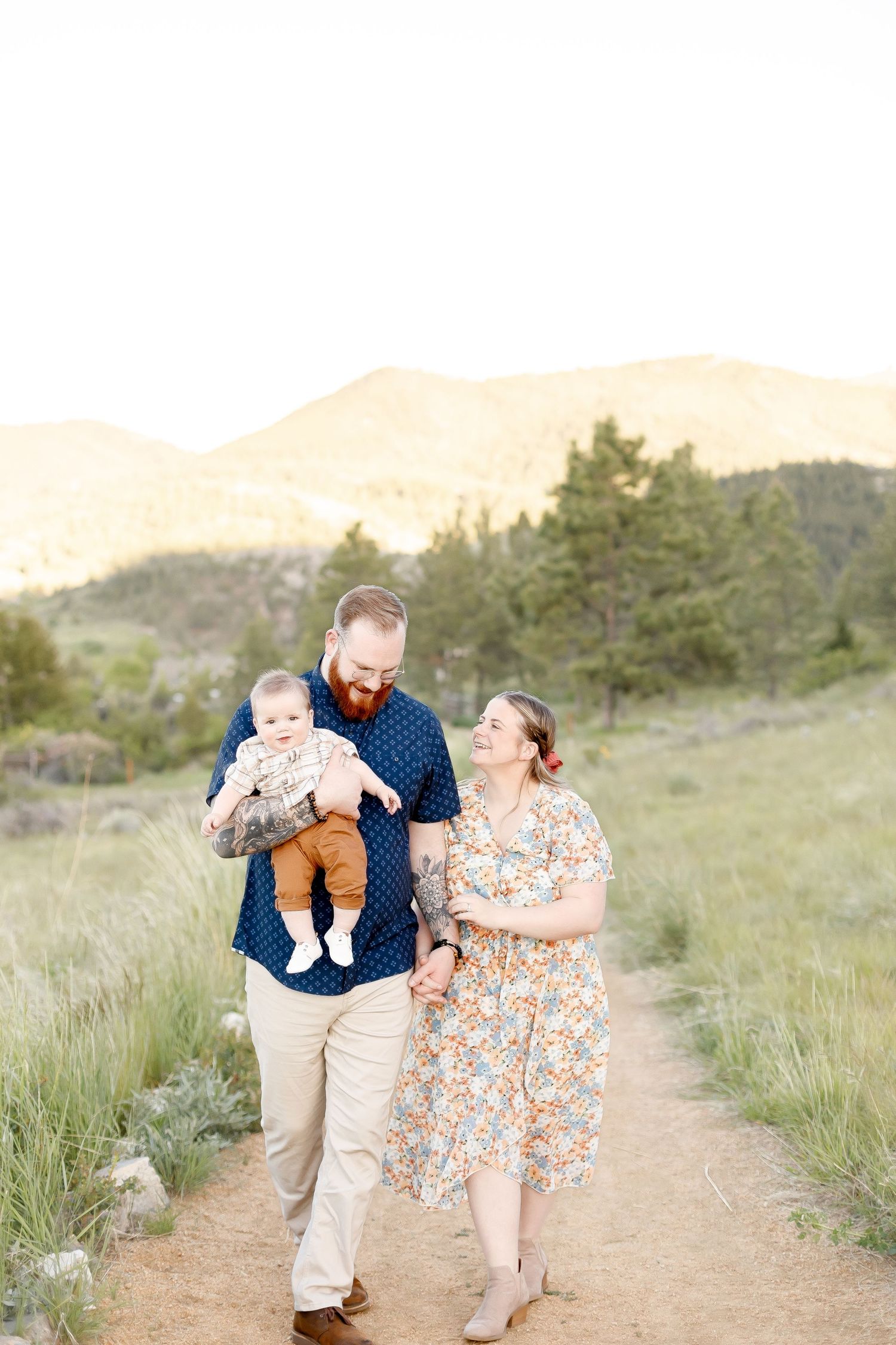 Young family with red-bearded father holding baby walks on mountain trail path at golden sunset.