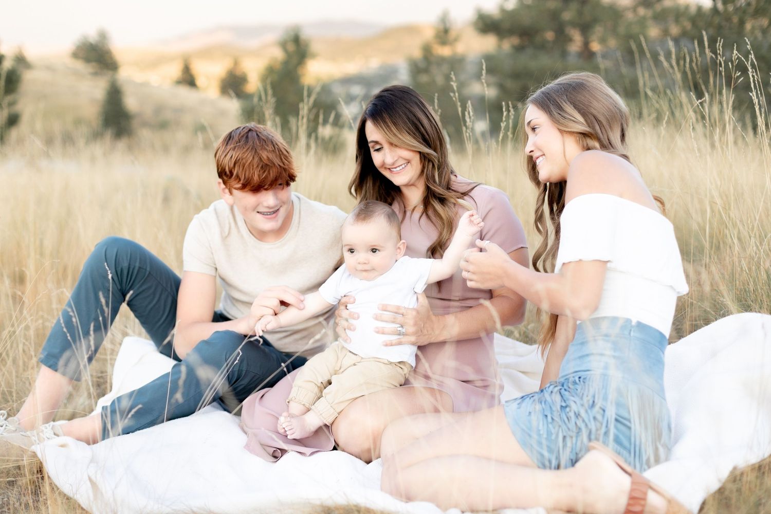 Three siblings sit on a blanket in a golden field, smiling and playing with a baby outdoors.