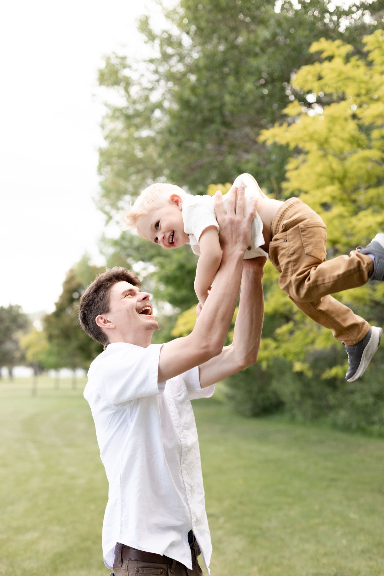 Laughing father lifting his happy toddler son high in the air in a sunny green park.