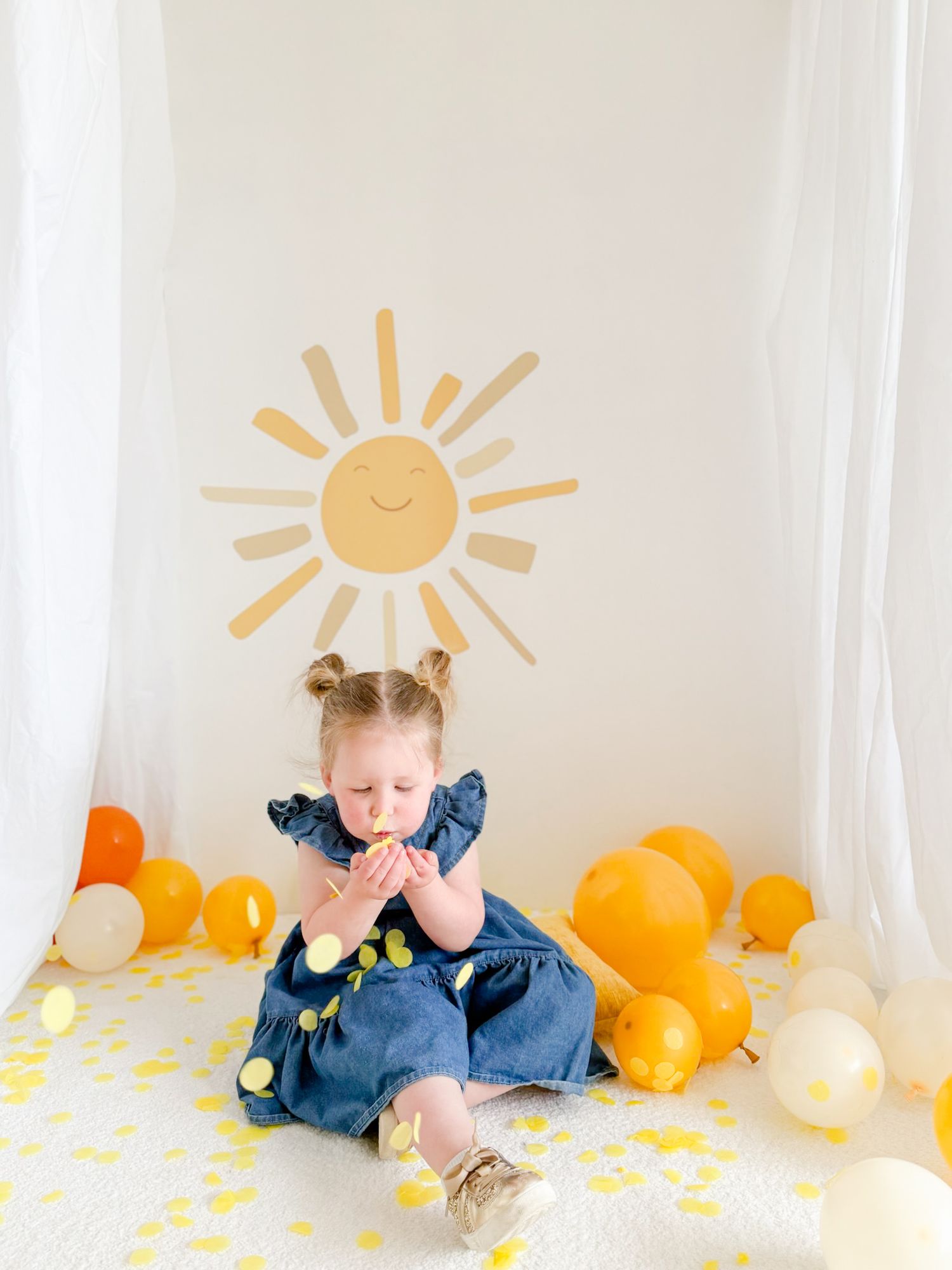 Toddler girl in denim dress sitting among orange and yellow balloons with sun wall decal.