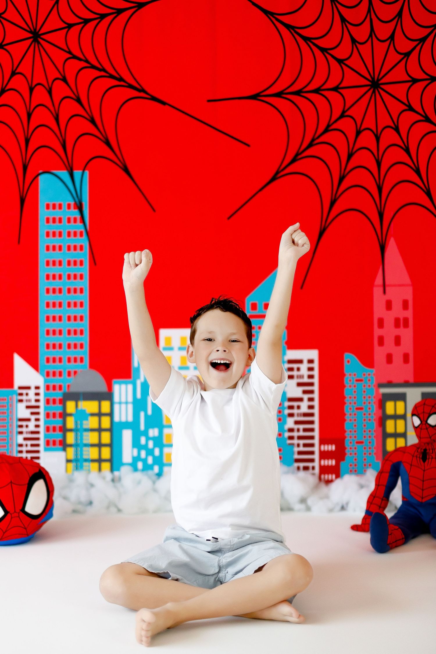 An excited boy raises his fists in celebration before a colorful Spider-Man themed birthday backdrop.