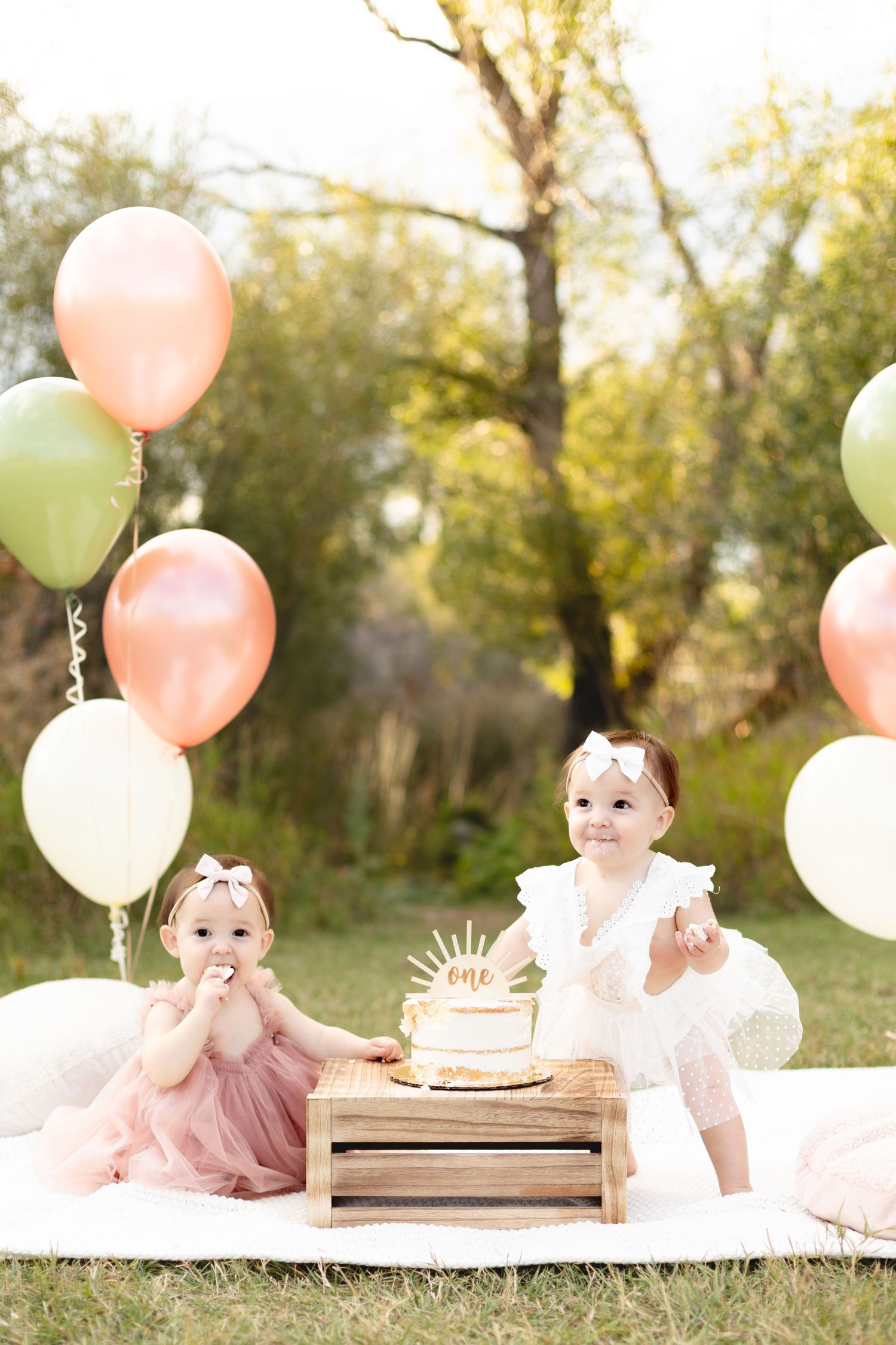 Twin baby girls celebrating first birthday with cake smash surrounded by pink and green balloons outdoors.