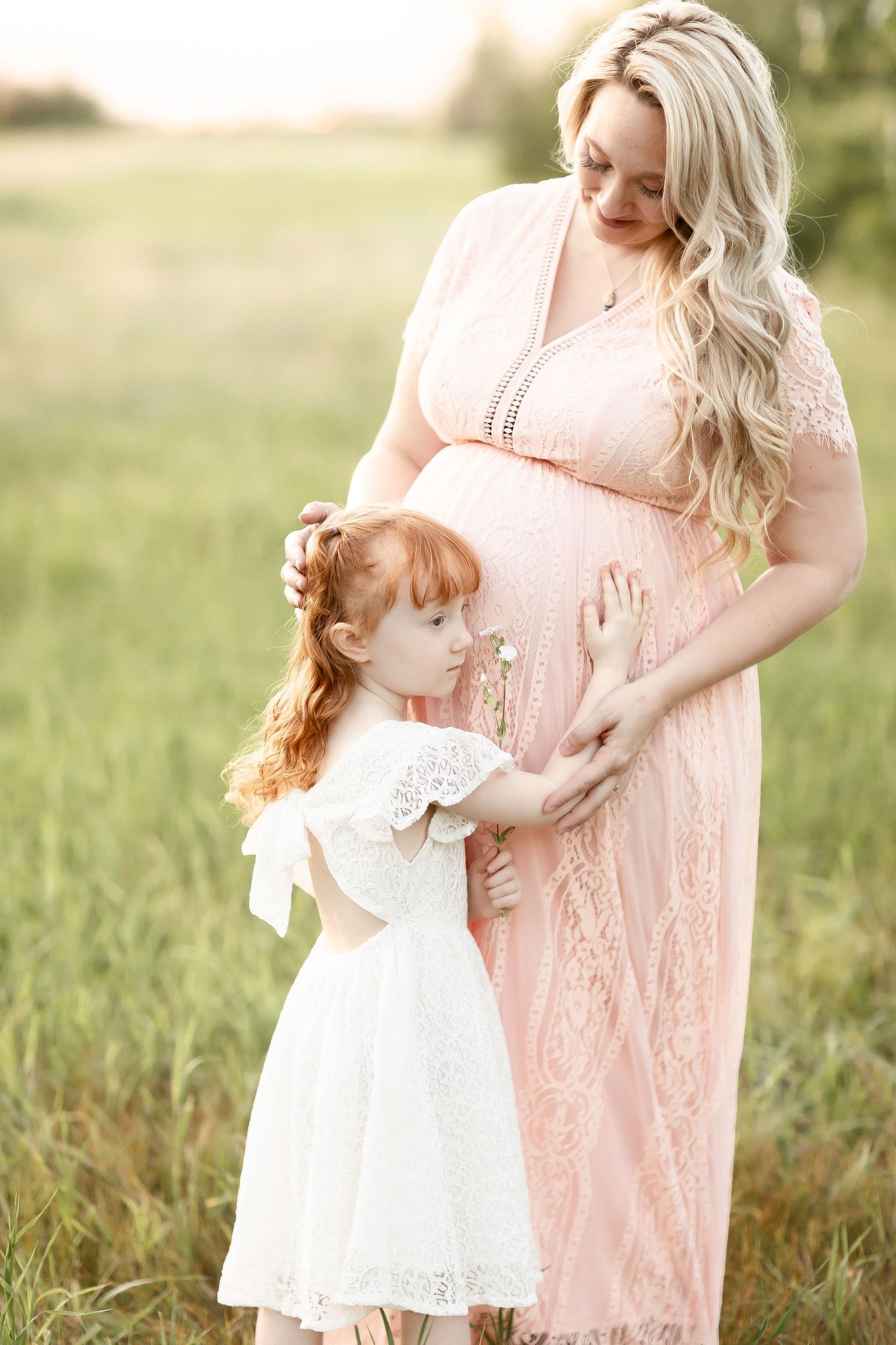 Little redhead girl touching pregnant mothers belly outdoors in golden hour light.
