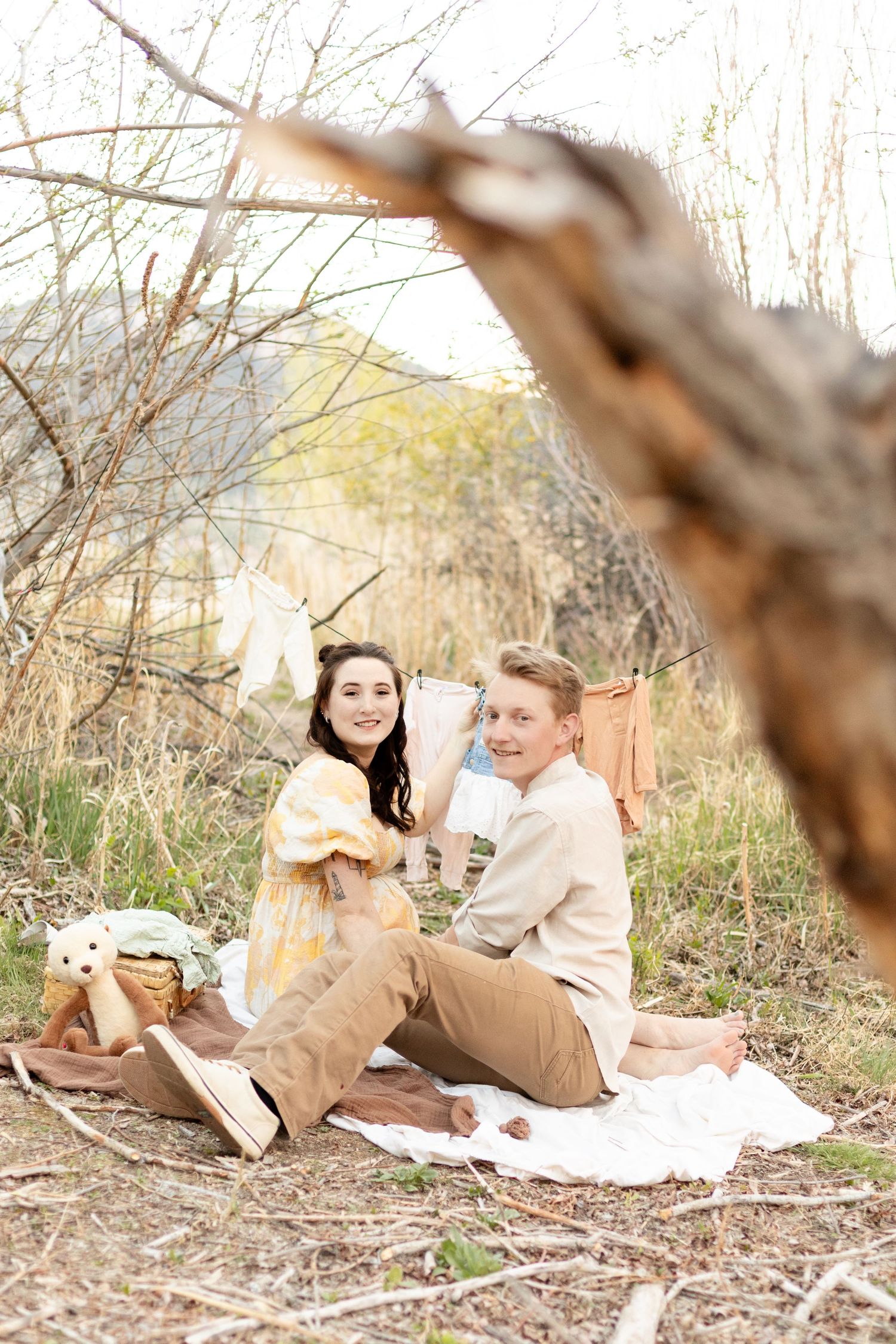 Smiling expecting couple sits on outdoor blanket holding baby clothes with a clothesline of infant items behind them.