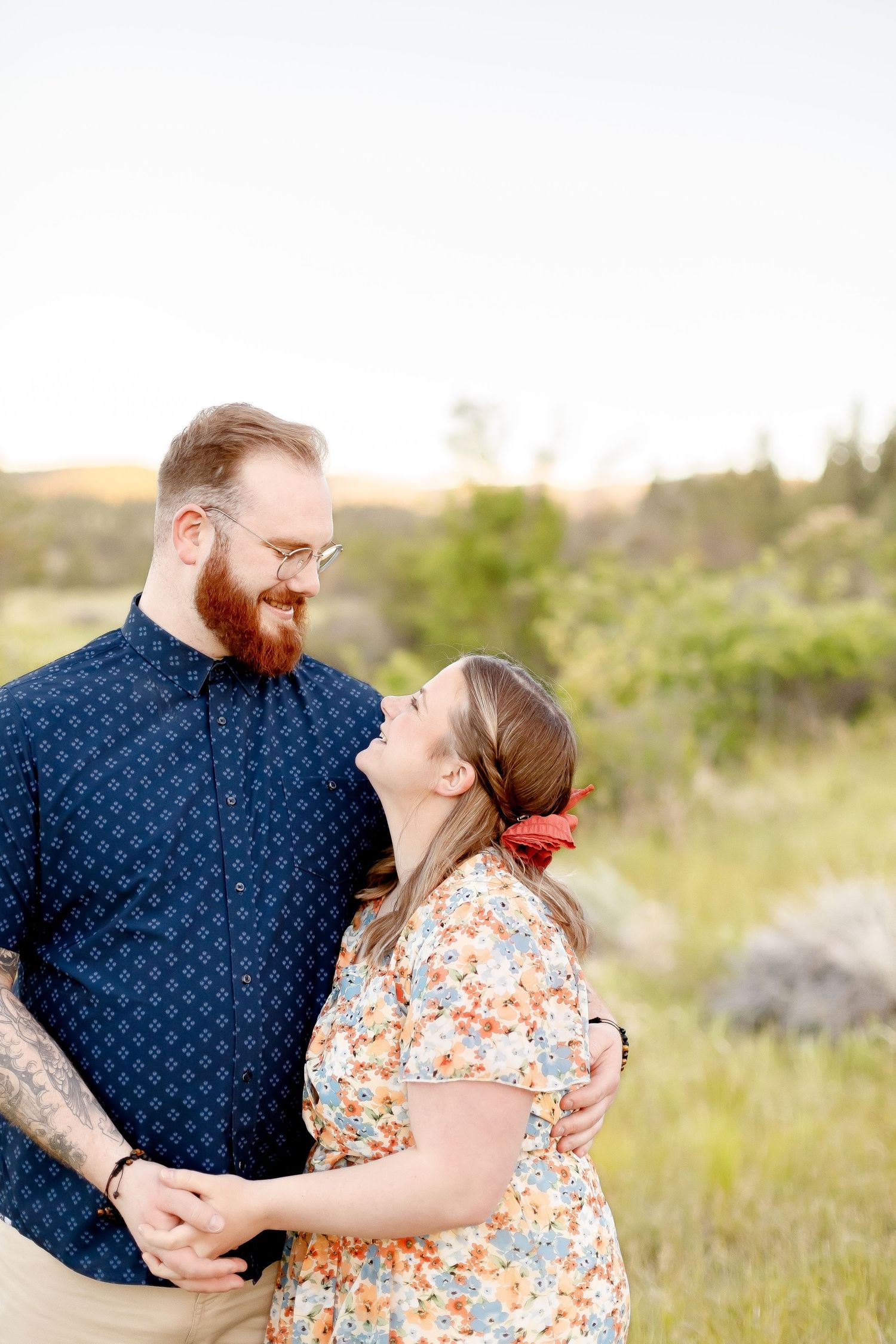 Bearded man in navy shirt and woman in floral dress laughing together in a sunlit green meadow.