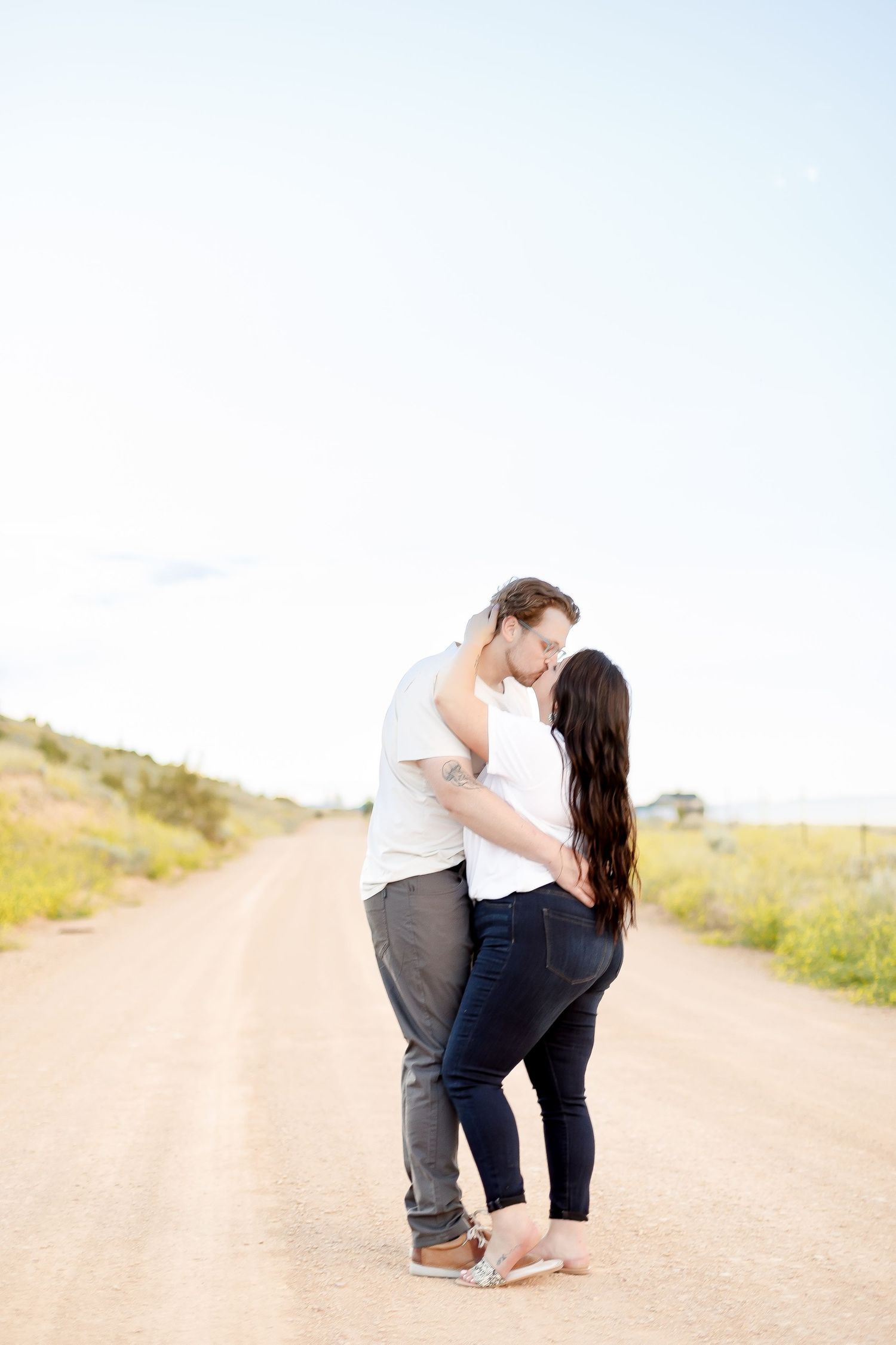 Couple sharing a romantic moment on a sunlit dirt road, woman in jeans, man in white shirt.