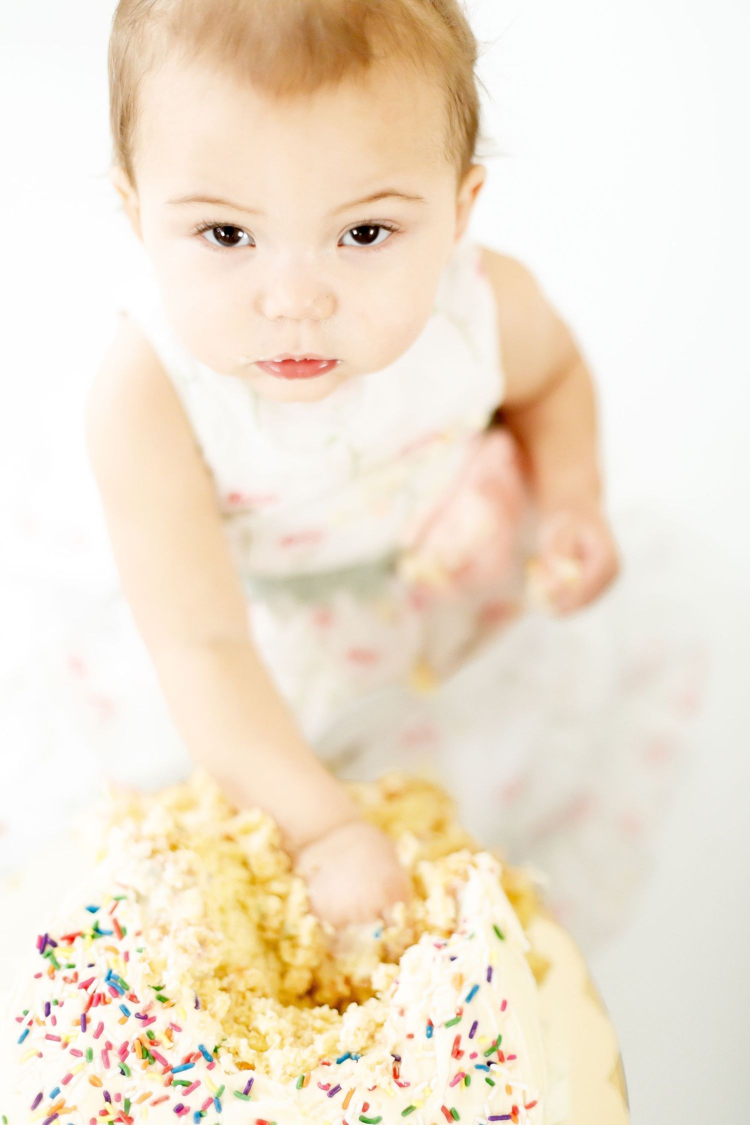 Baby girl in floral romper smashing white sprinkle cake during first birthday cake smash session.