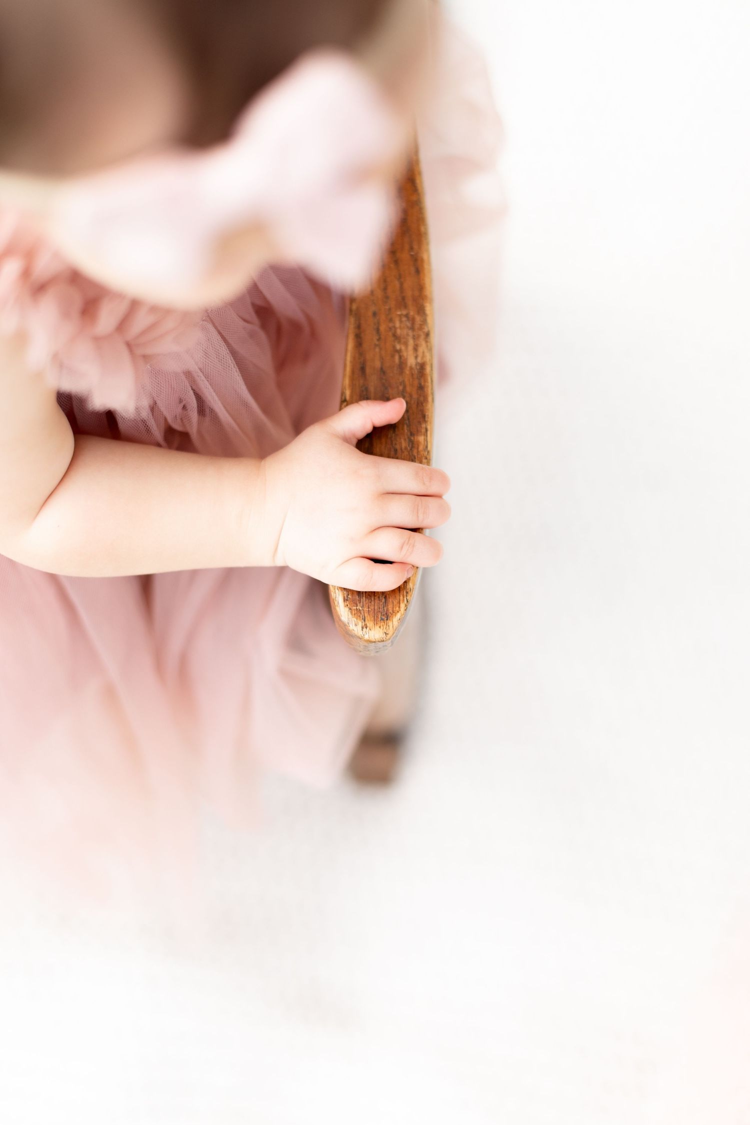 Close-up of baby girl in pink tulle dress holding wooden chair edge during first birthday photo session.