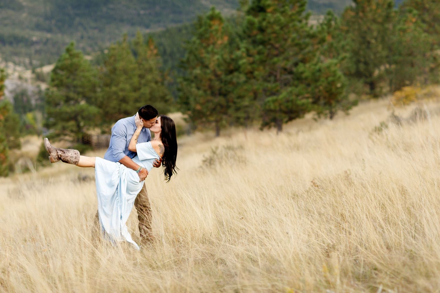 Couple sharing a romantic kiss in a golden field with pine trees and mountains in the background.