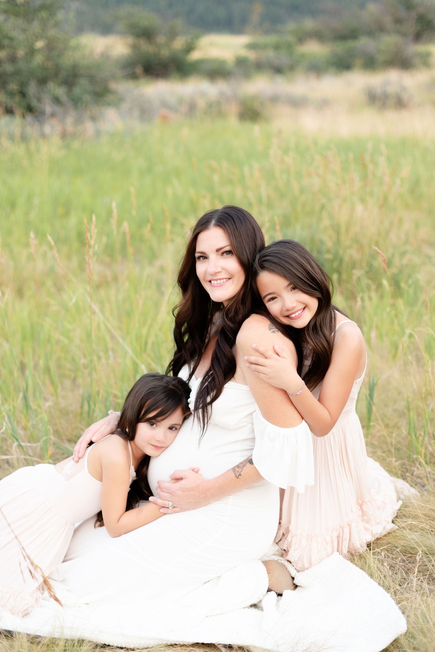 Pregnant woman sitting in field with two daughters hugging her during maternity session.