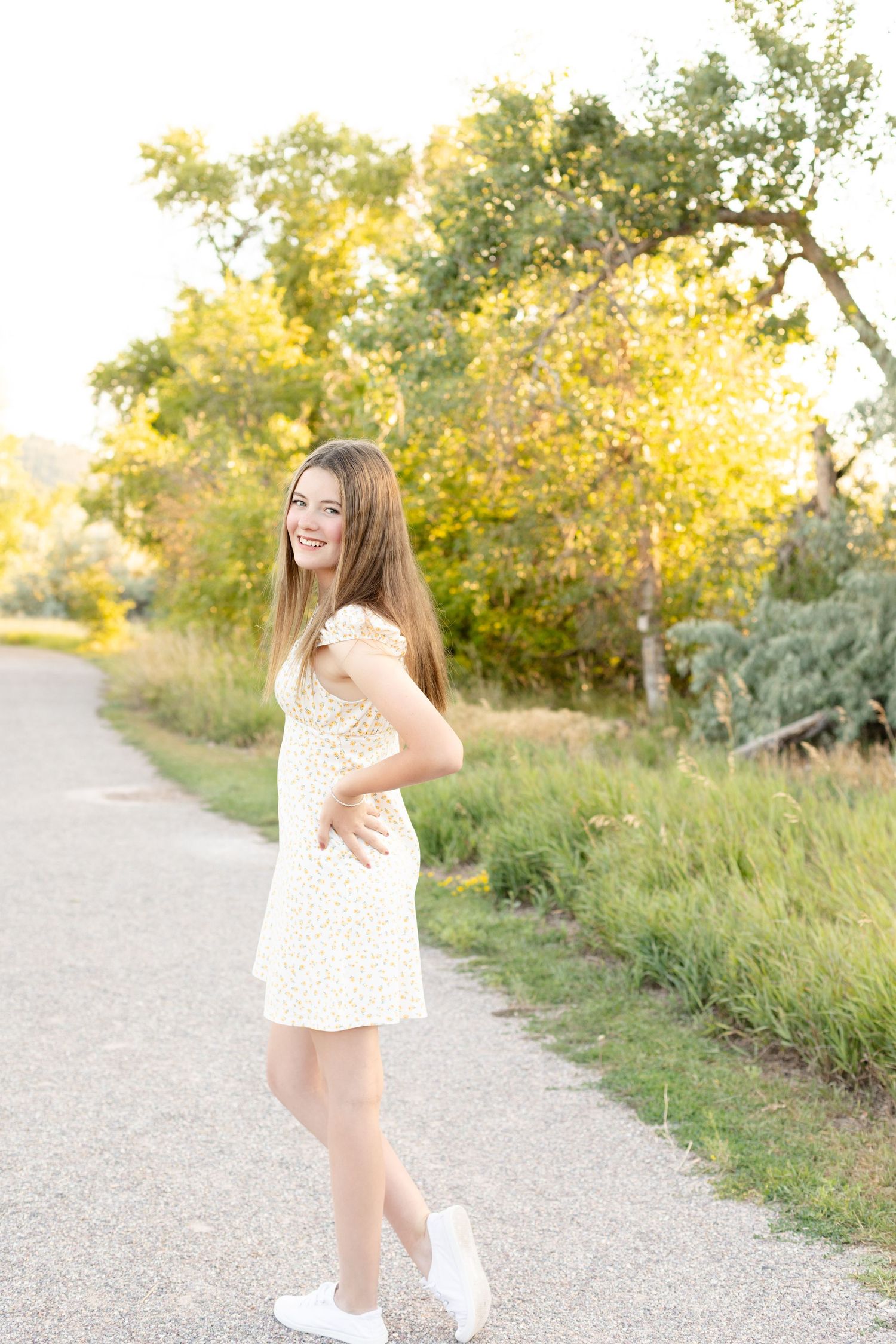 Happy young woman in yellow floral dress posing on a sunlit path surrounded by lush green foliage.