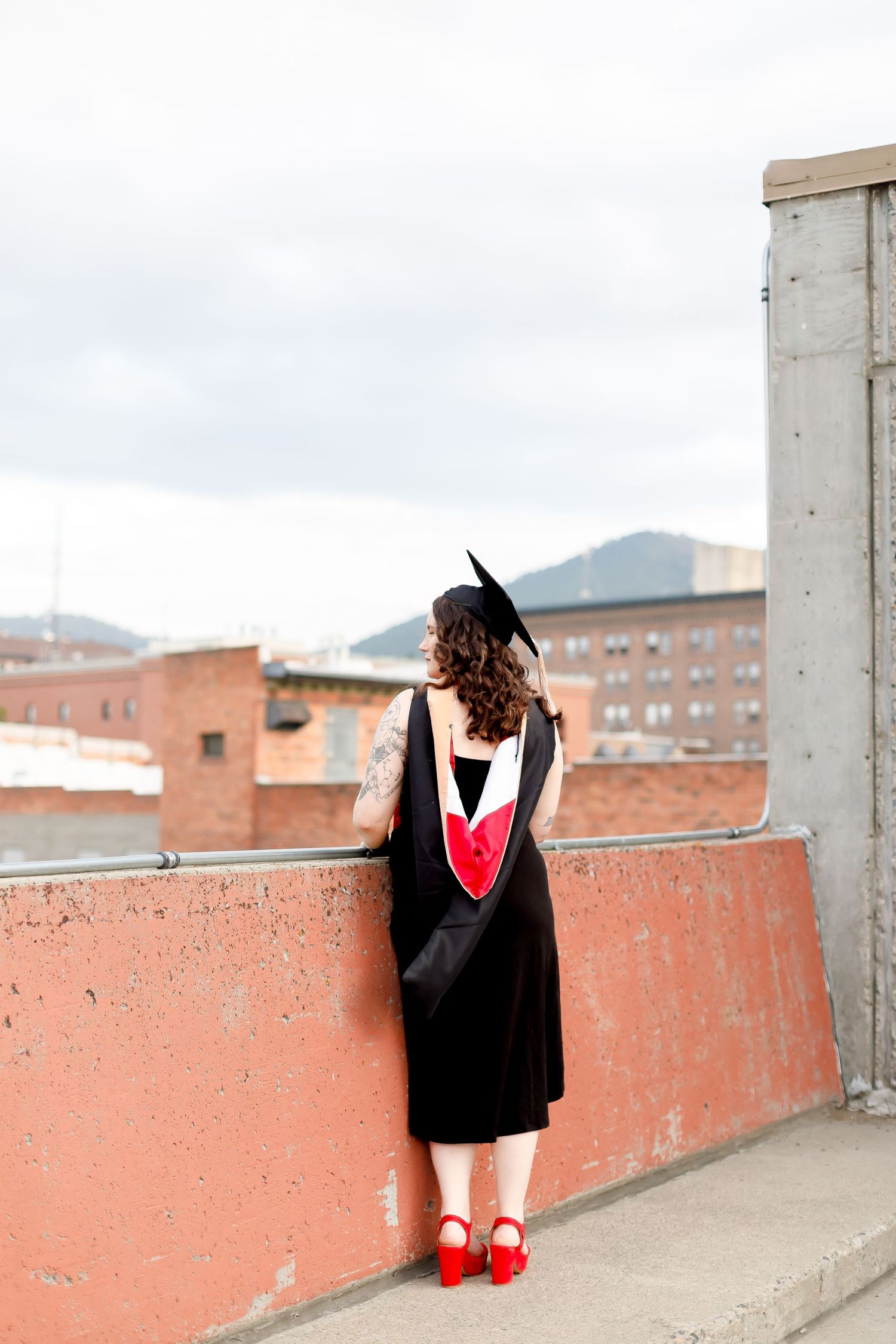 Graduate in black gown and red stole standing with back to camera overlooking a city rooftop view.